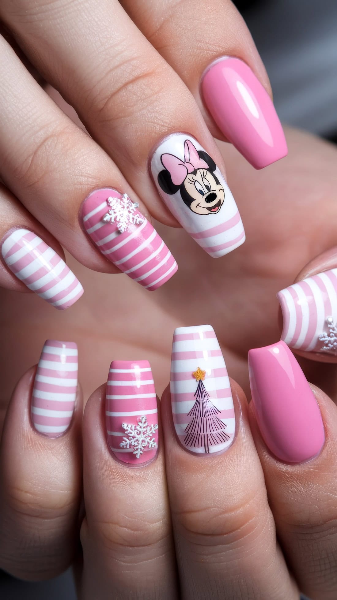 Close-up of hands with trendy Disney Christmas nails. Pink and white stripes adorn most, while one nail showcases Minnie Mouse with a bow, another displays a delicate snowflake, and a third features a purple Christmas tree topped with a shimmering gold star.