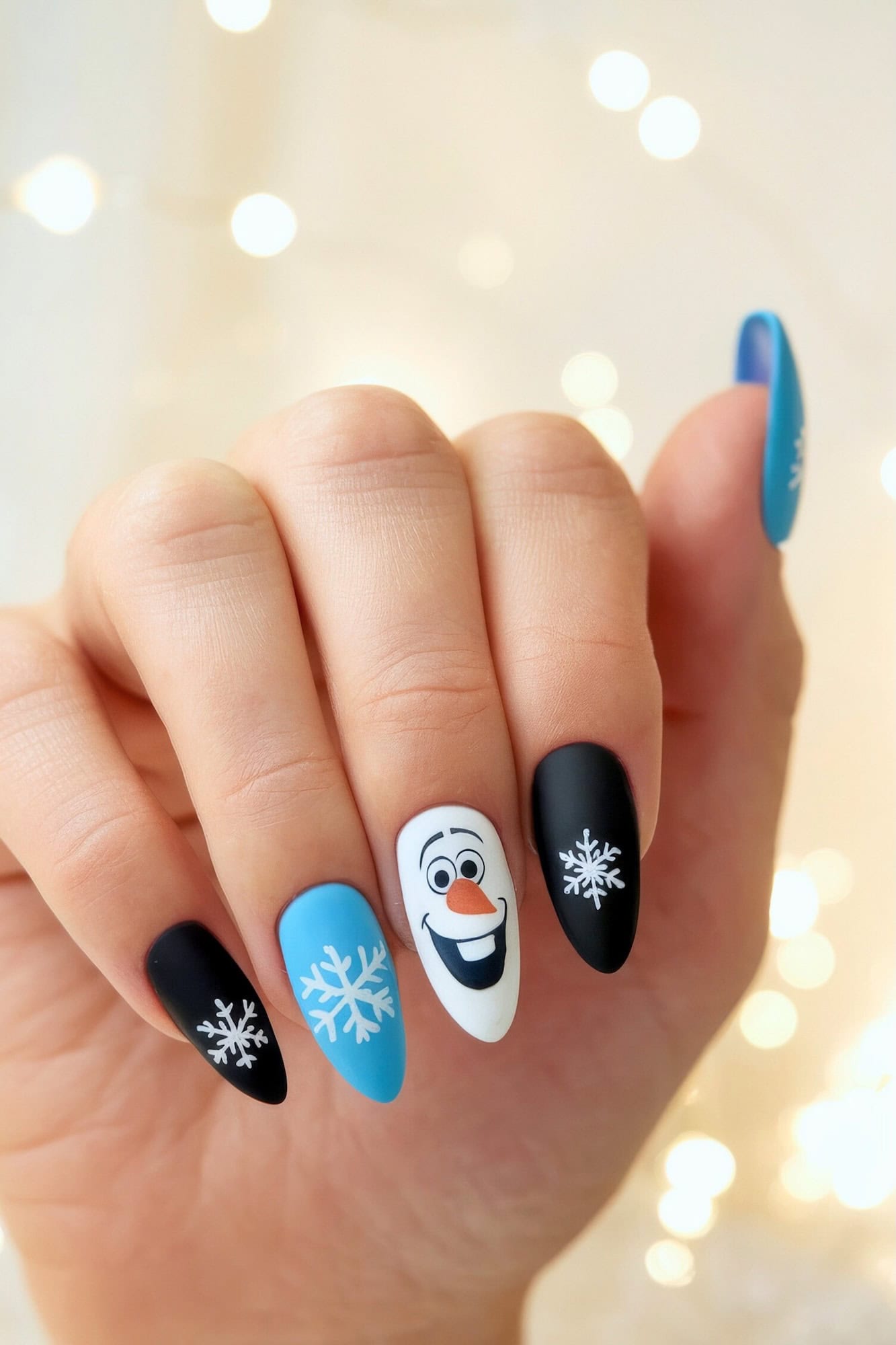 A hand showcases Disney Christmas nails with winter-themed art. The black, blue, and white nails feature snowflakes and a smiling snowman character, Olaf from Frozen. The background is softly blurred with warm, glowing lights.