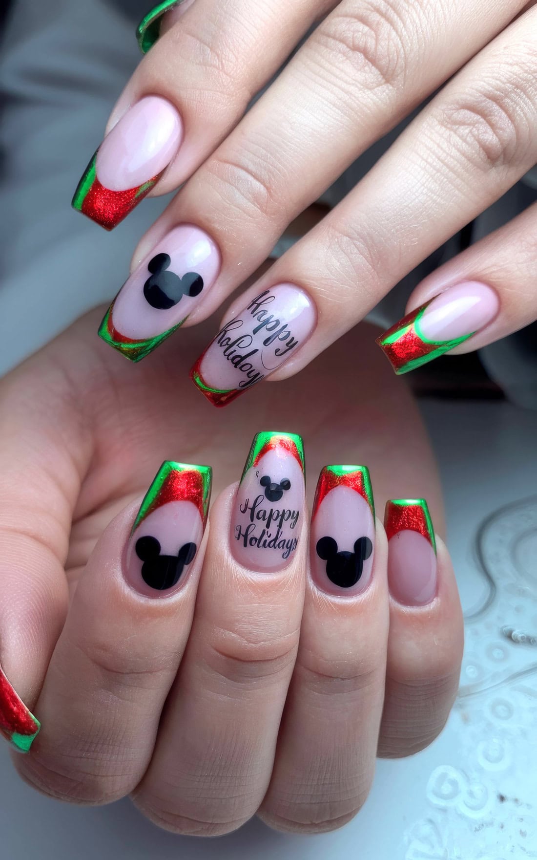 Close-up of hands with Disney Christmas nails featuring holiday-themed art. Nails showcase red and green French tips, playful Mickey Mouse ears silhouettes, and "Happy Holiday" text on the ring fingers. These festive nails capture a whimsical, Christmas spirit perfect for any Disney fan.