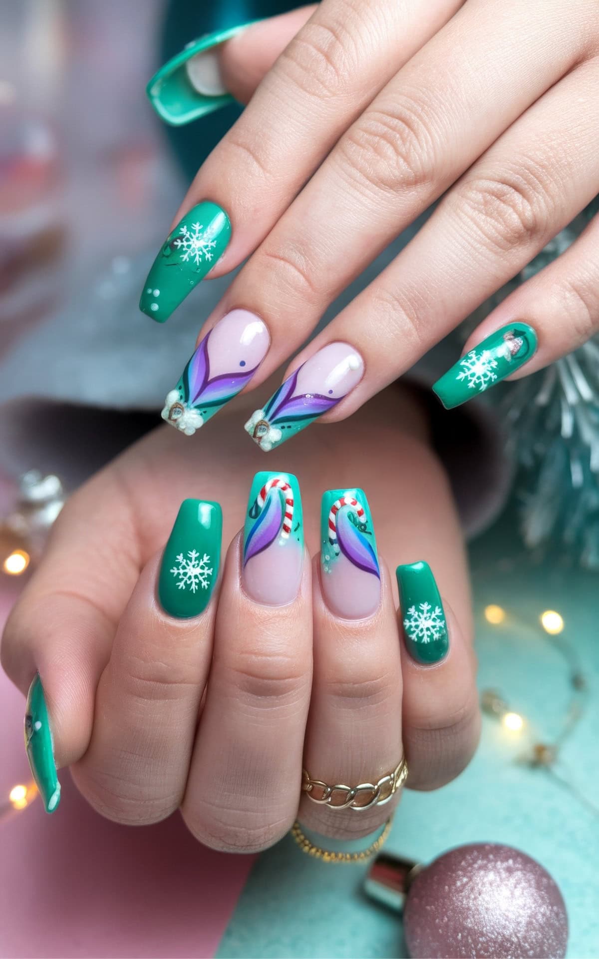 A close-up of manicured hands showcases festive Disney Christmas nails featuring princess Ariel from The Little Mermaid. Painted green, the nails feature snowflakes, swirls, and gem accents with playful Disney and mermaid flair. The background is adorned with subtle holiday lights and a shiny silver ornament, creating a cheerful, seasonal vibe.