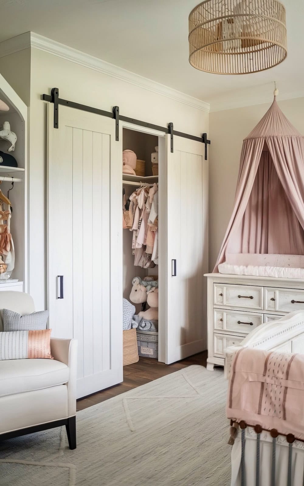 Cozy farmhouse nursery decor featuring a cream-colored armchair, a white dresser, and a crib draped with a pink canopy. The sliding closet doors reveal neatly arranged baby clothes and stuffed animals. A woven light fixture adds rustic charm from the ceiling.