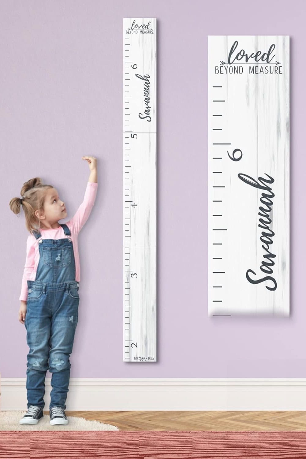 A young girl stands next to a large vertical growth chart in charming farmhouse nursery decor. She reaches up excitedly with one hand. The chart features the name "Savannah" and the phrase "Loved Beyond Measure" in decorative script on a light purple wall.