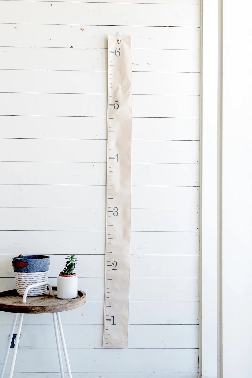 A white growth chart hangs on a white shiplap wall, marked with numbers 1 through 6, embodying farmhouse nursery decor. Next to it, a small table holds a blue basket and a potted succulent plant.