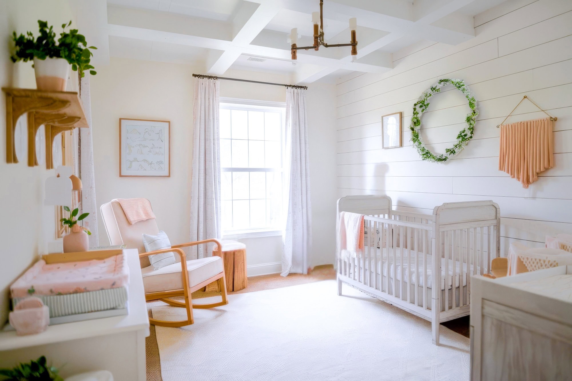 A bright and airy farmhouse nursery with white walls and a large window embraces farmhouse nursery decor. It features a white crib, a rocking chair with a pink cushion, a changing table, and walls adorned with a wreath and macramé hanging. Soft, natural lighting fills the room.
