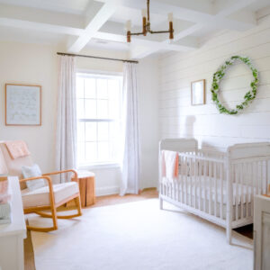 A bright, airy nursery showcases farmhouse decor with a white crib, a wooden rocking chair draped in a peach blanket, and a round green wreath on the wall. Sunlight pours through a large window with light curtains, illuminating the room's white tones and rustic wooden accents.