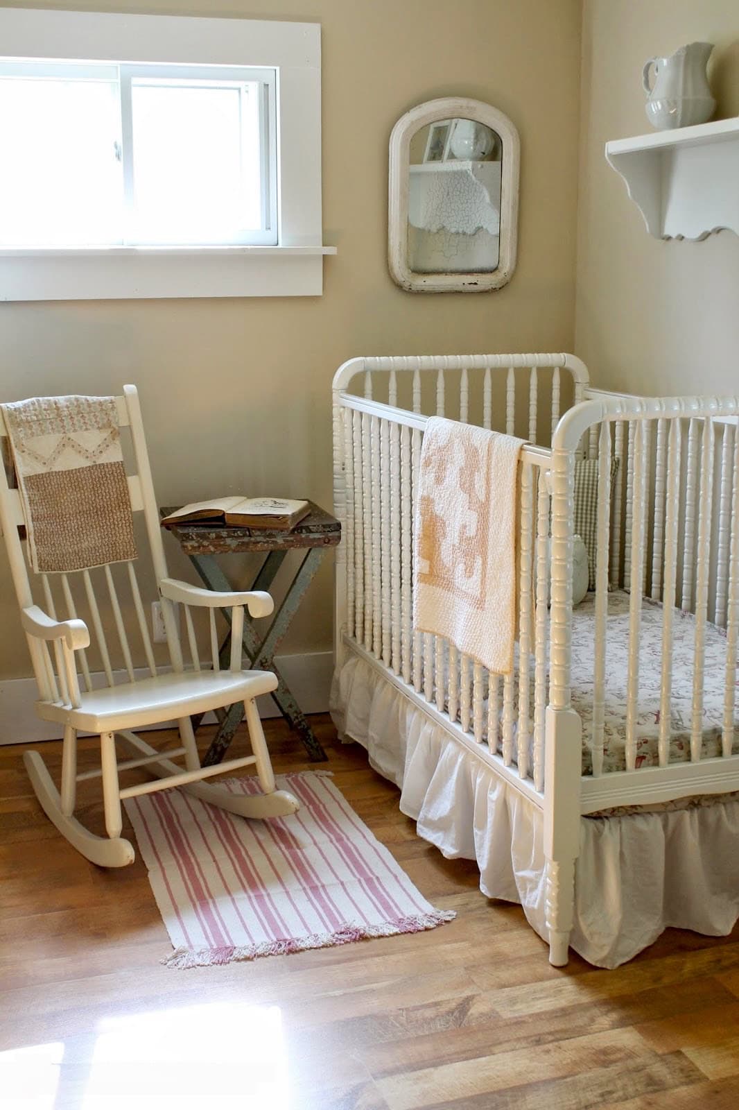 A cozy nursery features farmhouse decor with a white wooden crib adorned with a quilt next to a light-colored rocking chair. A small side table holds a decor item, with a mirror above. A striped rug covers part of the wooden floor, while sunlight streams through the window.