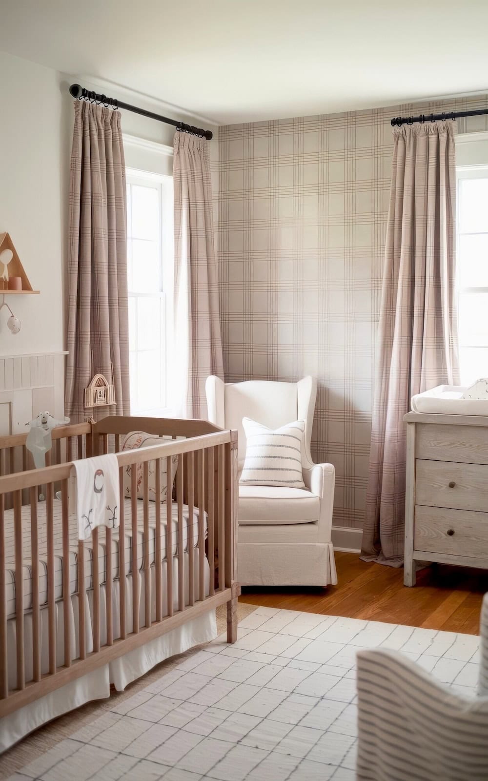 A cozy farmhouse nursery with plaid wallpaper, a wooden crib, and a white cushioned rocking chair. The room features a wooden dresser and soft plaid curtains on large windows, with light wood flooring and a patterned rug adding to the warm ambiance.