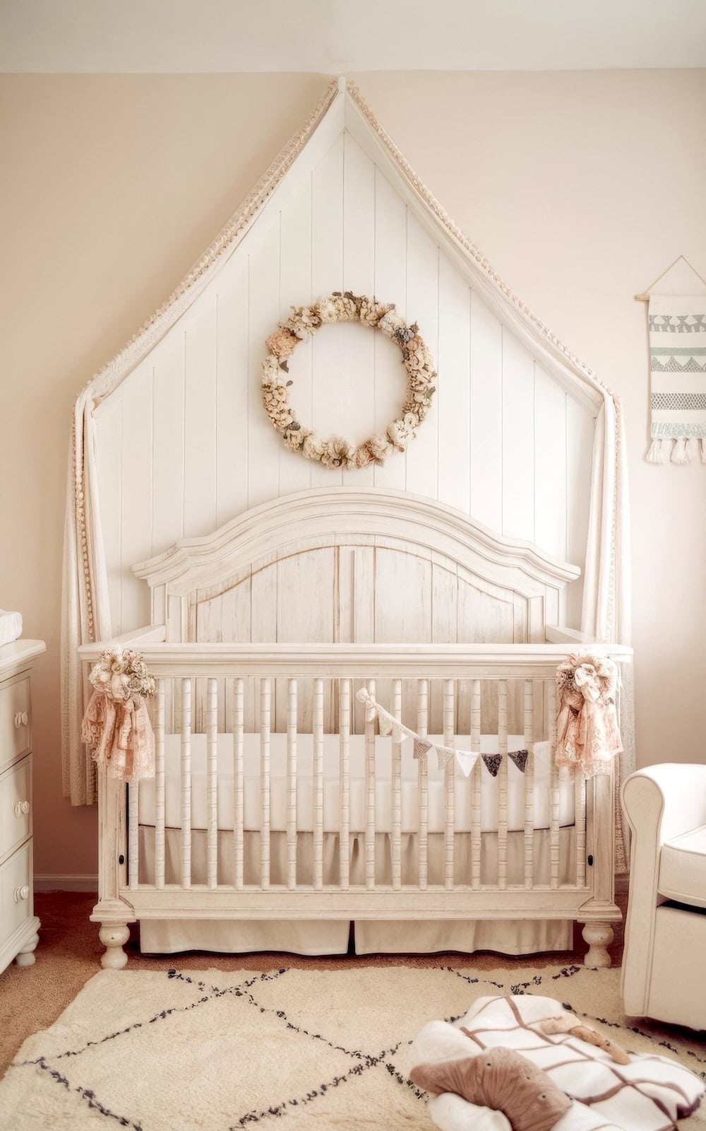 A cozy nursery showcases farmhouse decor with a wooden crib adorned with floral decorations and a small pennant banner. There's a cutout section of shiplap behind the crib. There's a delicate wreath on a pointed backdrop, soft lighting, a comfy chair on the right, and a patterned rug on the floor.