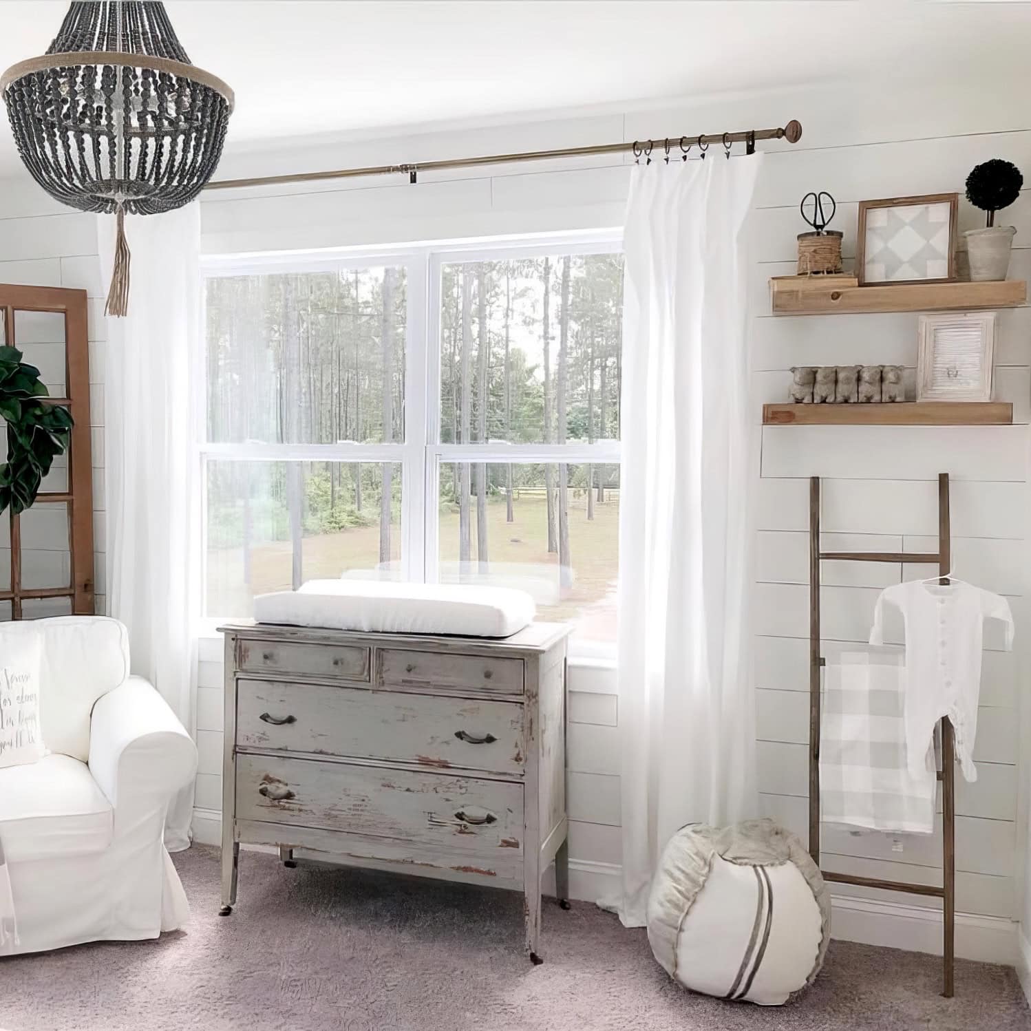 A bright farmhouse nursery decor features a large window and a vintage vanity repurposed as a changing table. A white armchair, decorative ladder with blanket, and shelves adorned with small decor complete the room. A beaded chandelier elegantly hangs from the ceiling.