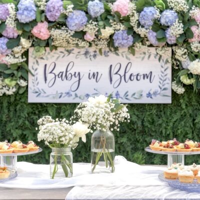 A spring baby shower setup with a sign reading "Baby in Bloom" surrounded by vibrant flowers, embracing spring baby shower themes. A white cloth-draped table holds vases of delicate white blooms and trays of small pastries, set against a backdrop of lush greenery with pink, blue, and purple blossoms.
