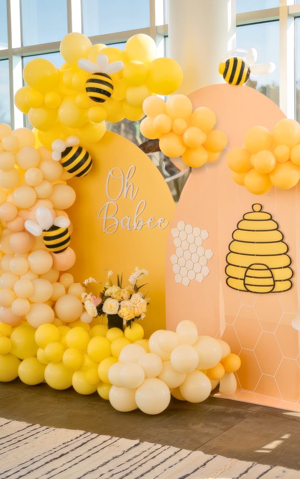 A decorative spring baby shower backdrop features yellow and white balloons arranged like clouds and bees. A beehive and honeycomb pattern display the words "Oh Babee" in the center, with yellow flowers blooming at the base, capturing a vibrant seasonal theme.