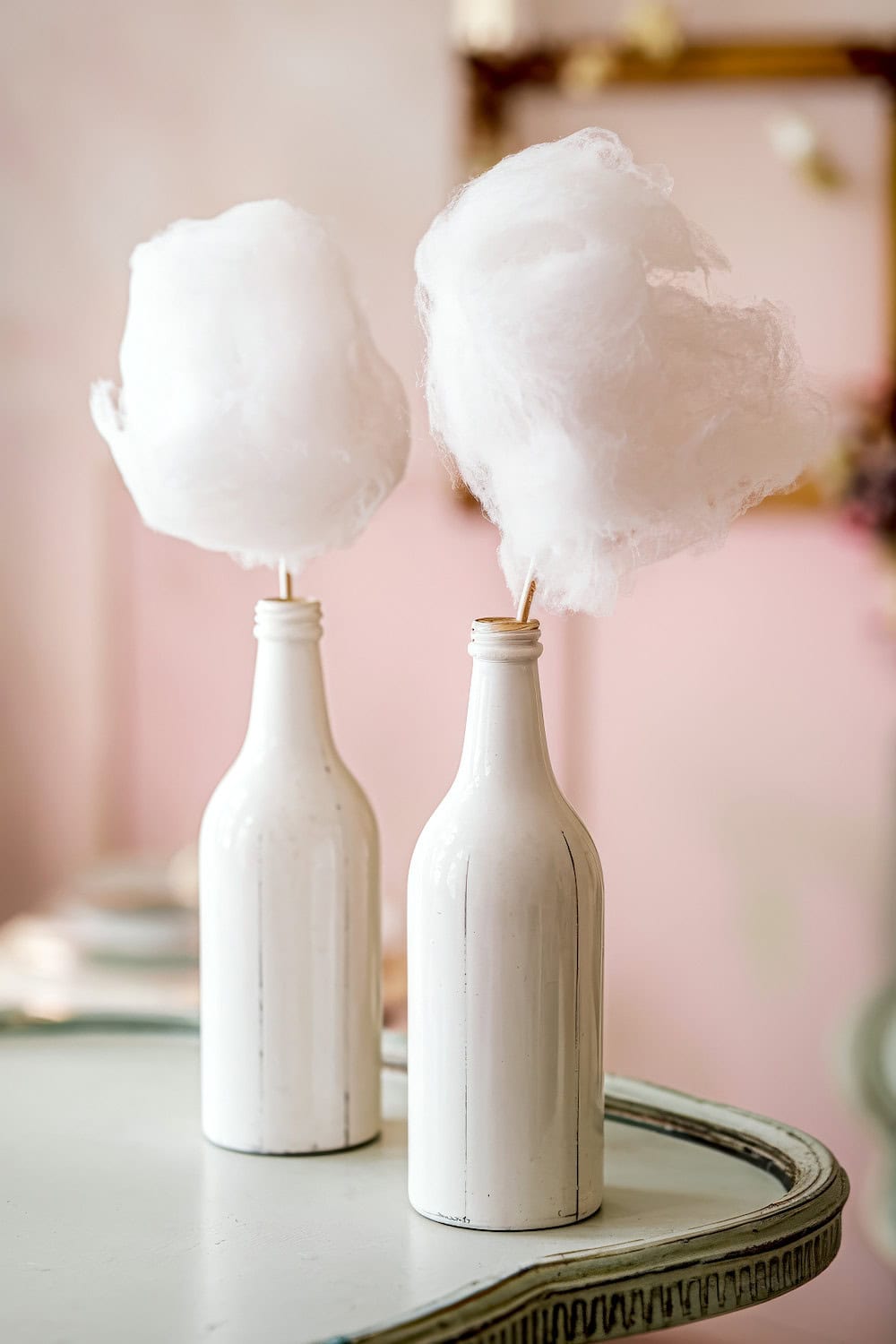 Two white bottles stand on a table, each holding a stick topped with fluffy white cotton candy, resembling clouds—a delightful nod to  Cloud 9 spring baby shower themes. The background is softly blurred with a pastel pink hue.
