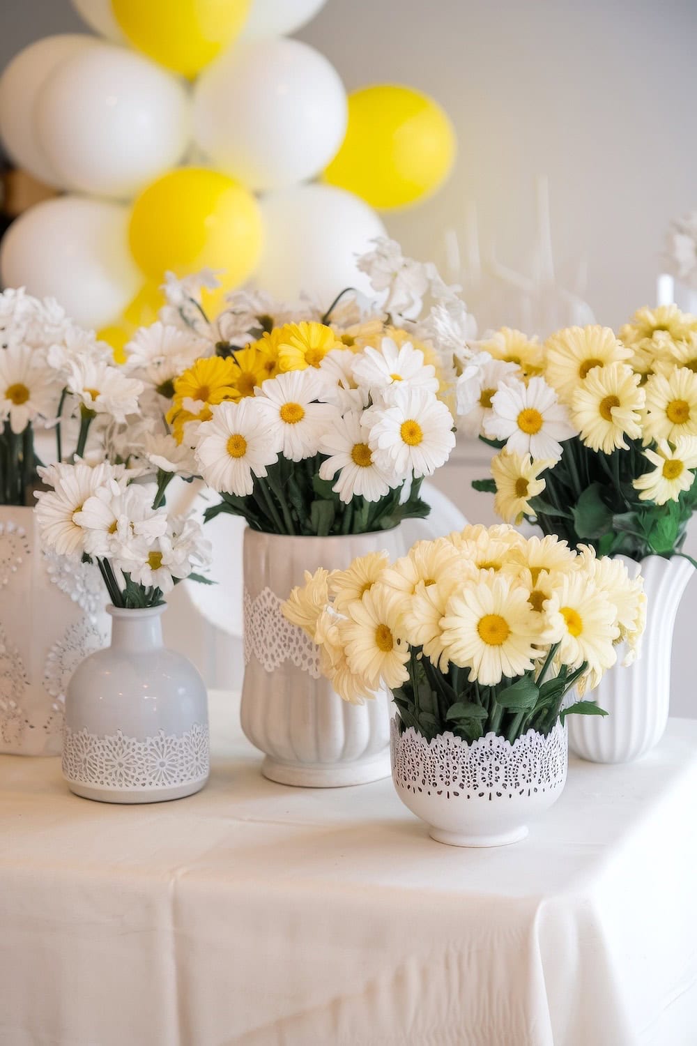 Vases filled with white and yellow daisies adorn the white table, perfect for spring baby shower themes. Decorative lace wraps around some of the vases, while blurred yellow and white balloons float in the background.