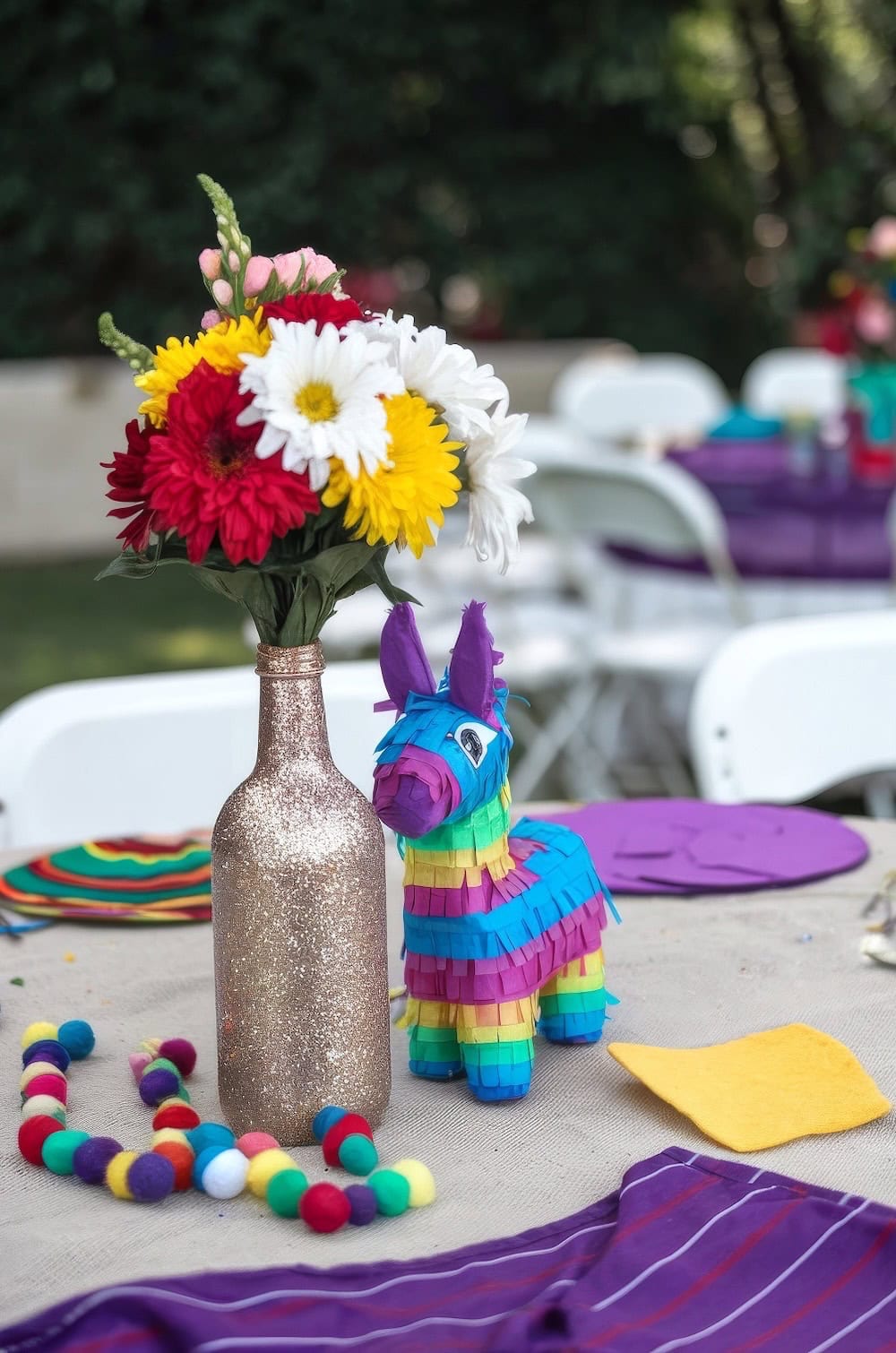 A festive fiesta baby shower outdoor table setting features a colorful donkey pinata, a glittery vase with vibrant flowers, and multicolored pom-poms, perfect for spring baby shower themes. The tablecloth is adorned with bright napkins and striped patterns, while white chairs surround the beautiful setup.