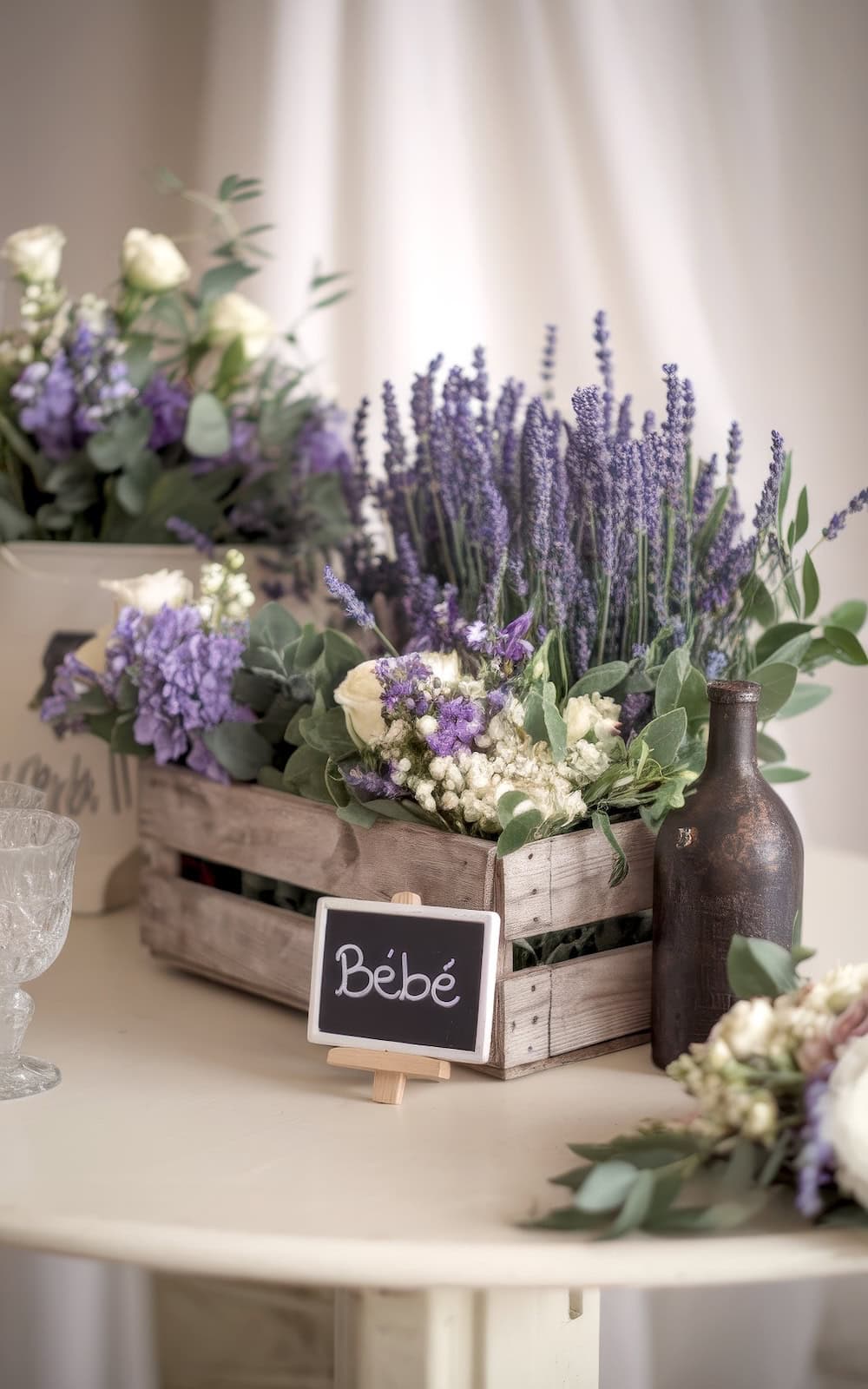 A wooden crate brimming with lavender and white flowers, labeled "Bébé" on a chalkboard, captures the essence of spring baby shower themes. A small brown bottle and crystal glass sit elegantly nearby, with additional flower arrangements enhancing the ambiance in the background.