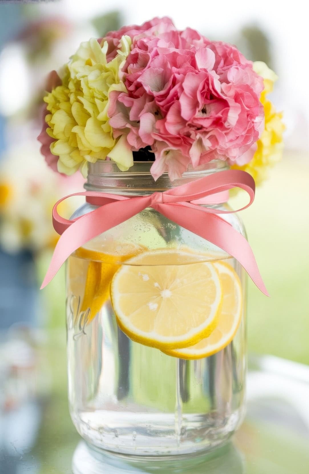 A glass jar filled with water and lemon slices, adorned with a pink ribbon, perfectly captures spring baby shower themes. Vibrant pink and yellow hydrangeas sit on top, creating a fresh and colorful arrangement for a lemonade baby shower against a softly blurred background.