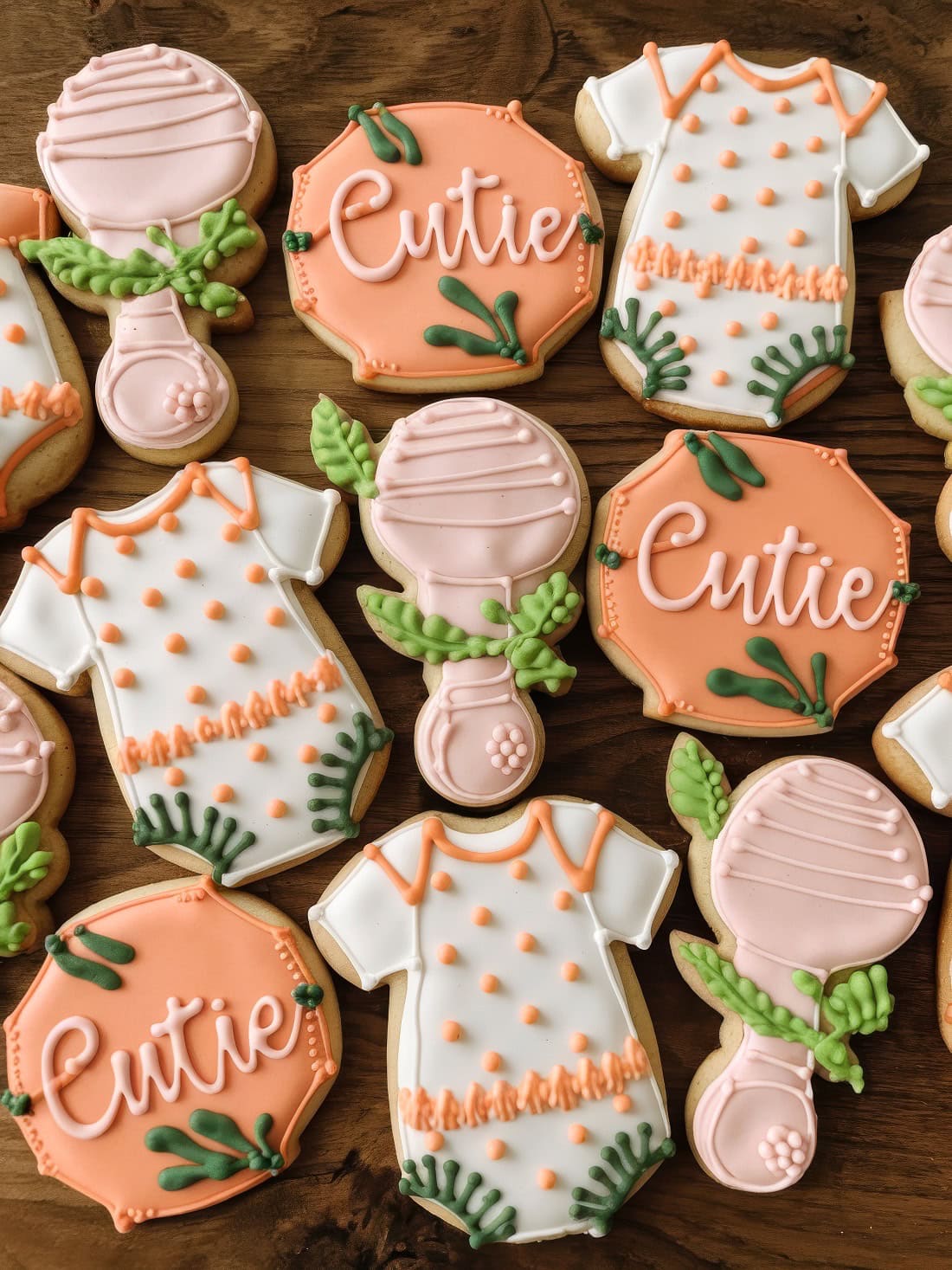 Decorated cookies for a Little Cutie baby shower on a wooden surface highlight delightful spring baby shower themes, featuring shirts, vases with flowers, and the word "Cutie" in orange and green accents with intricate icing details.