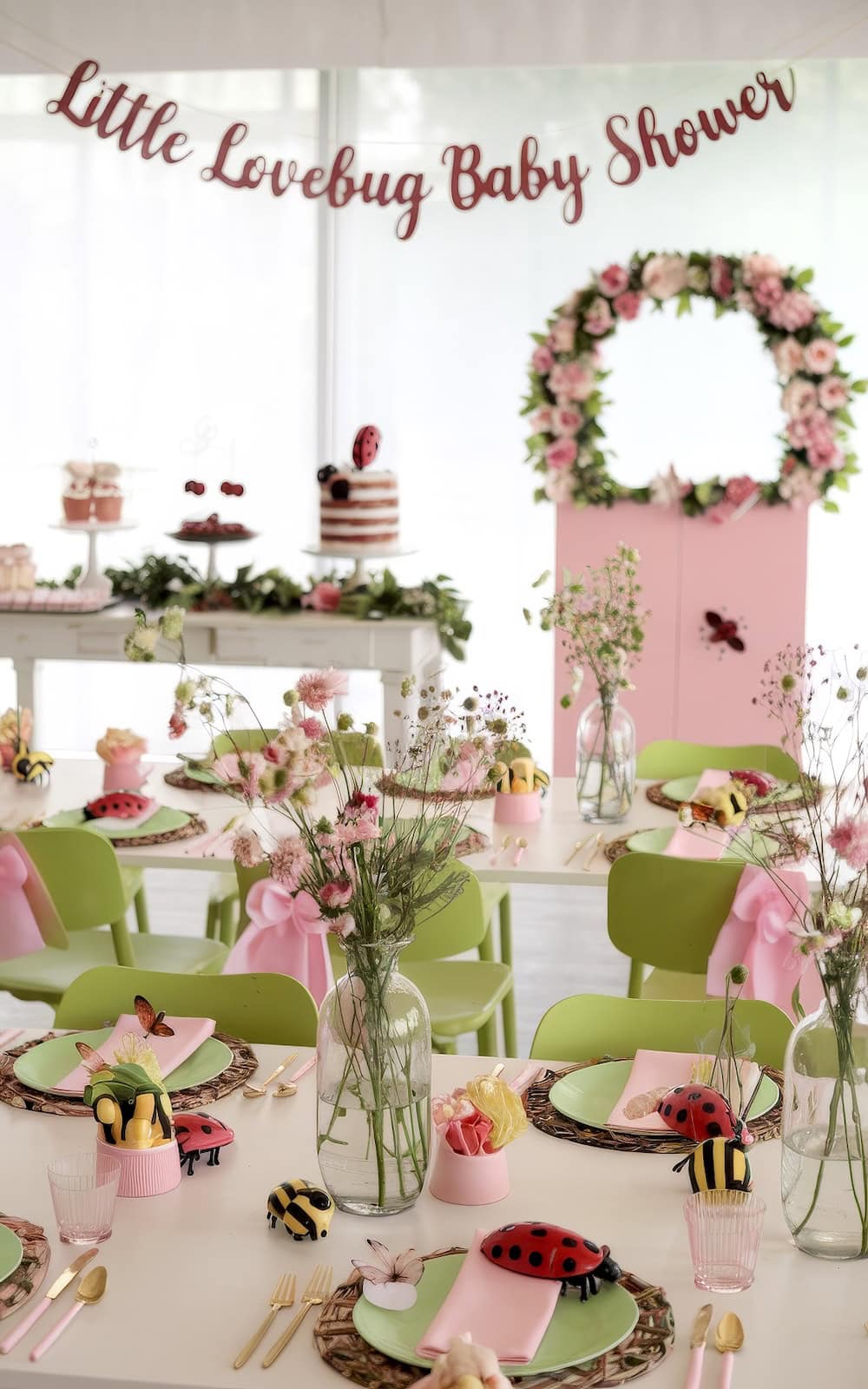 A baby shower table embraces spring baby shower themes with ladybug and bee plush toys, pink flowers, and green chairs. A banner reads "Little Lovebug Baby Shower" above a floral wreath. Cake and cupcakes patiently await on a side table.