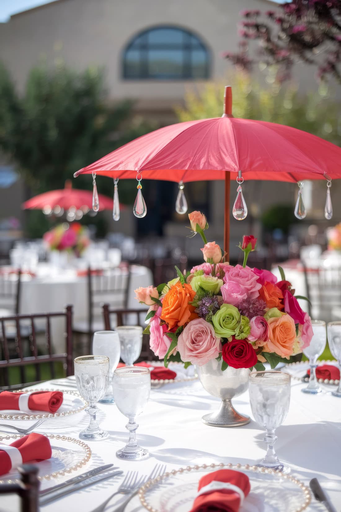 An outdoor spring baby shower event setup with a Raindrops on Roses theme. tables are covered in white cloth, each featuring a centerpiece of colorful flowers under a decorative red umbrella. Tables are set with glassware and red napkins, with a charming building and lush trees in the background.