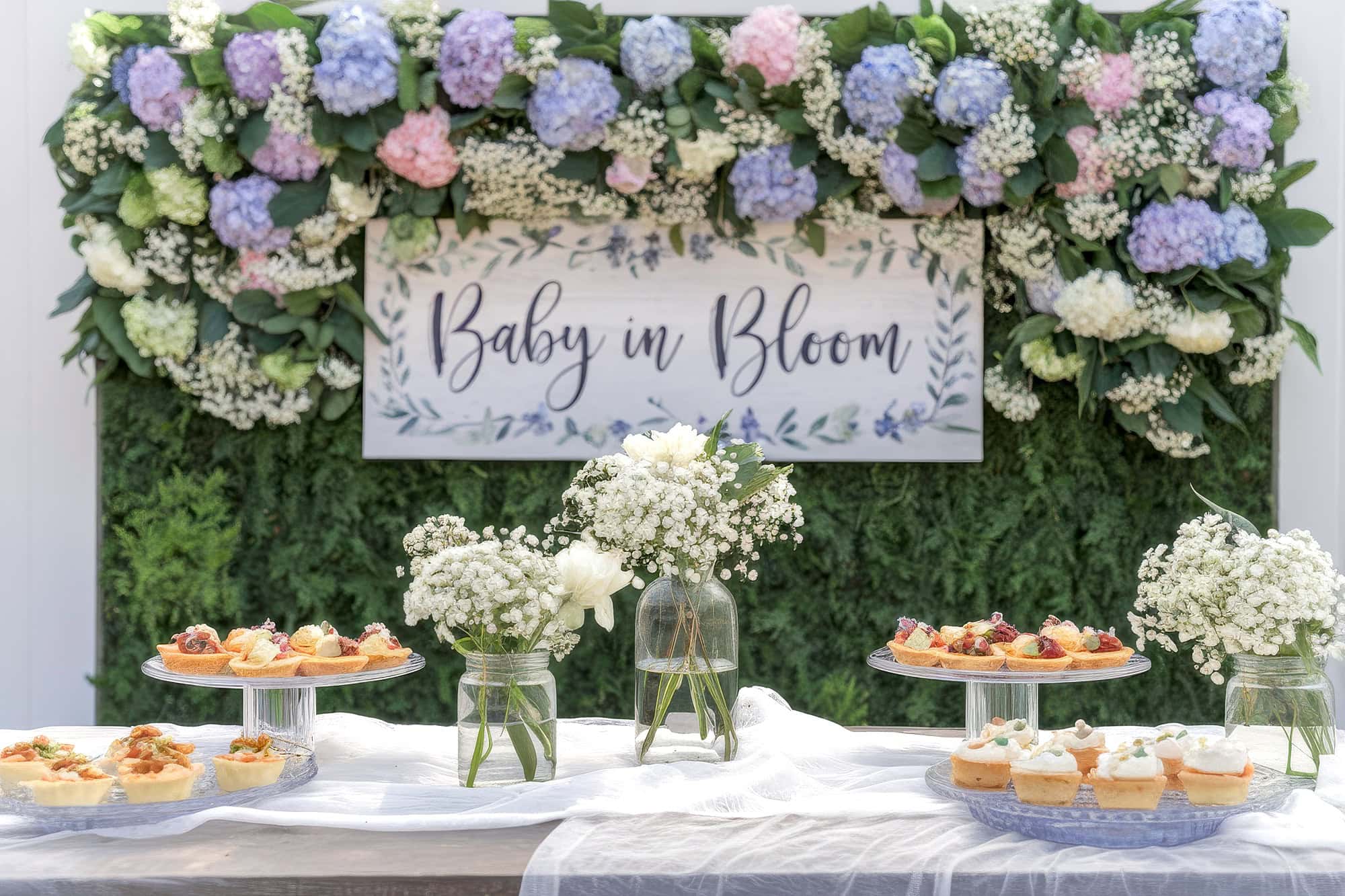 A decorative spring baby shower setup with a "Baby in Bloom" sign showcases spring baby shower themes. Floral arrangements of pastel hydrangeas and greenery abound, while a table is adorned with white flowers and tiered trays of assorted pastries.