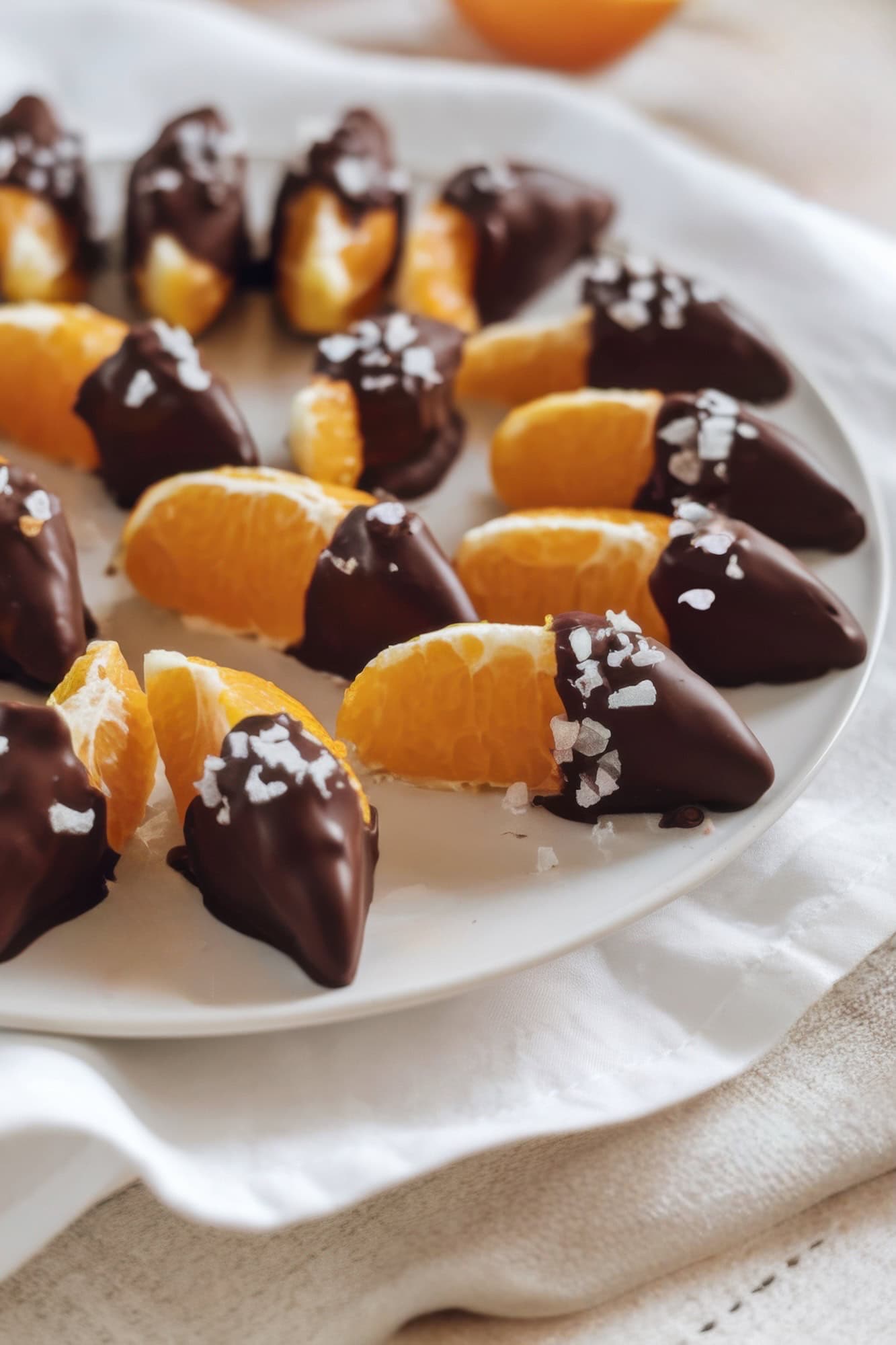 Slices of orange are partially dipped in dark chocolate and arranged in a circular pattern on a white plate, perfect for little cutie-themed baby shower ideas. The chocolate is sprinkled with coarse sea salt, and the plate rests on a white and beige fabric background.
