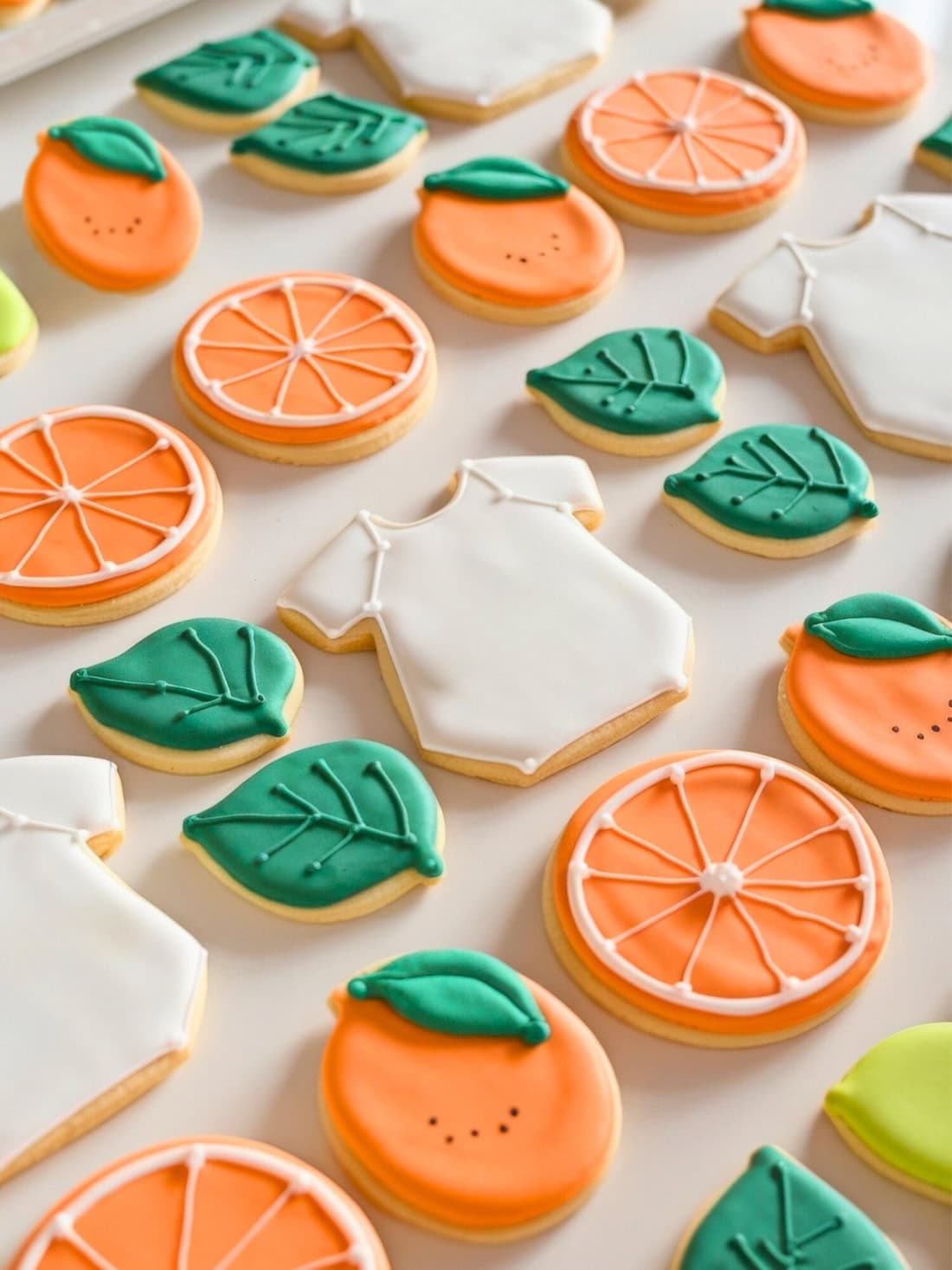 Assorted sugar cookies, perfect for little cutie baby shower ideas, are decorated with colorful icing: round cookies with orange slices, oranges with green leaves, green leaf-shaped cookies, and shirt-shaped cookies with white icing. All beautifully arranged on a white surface.