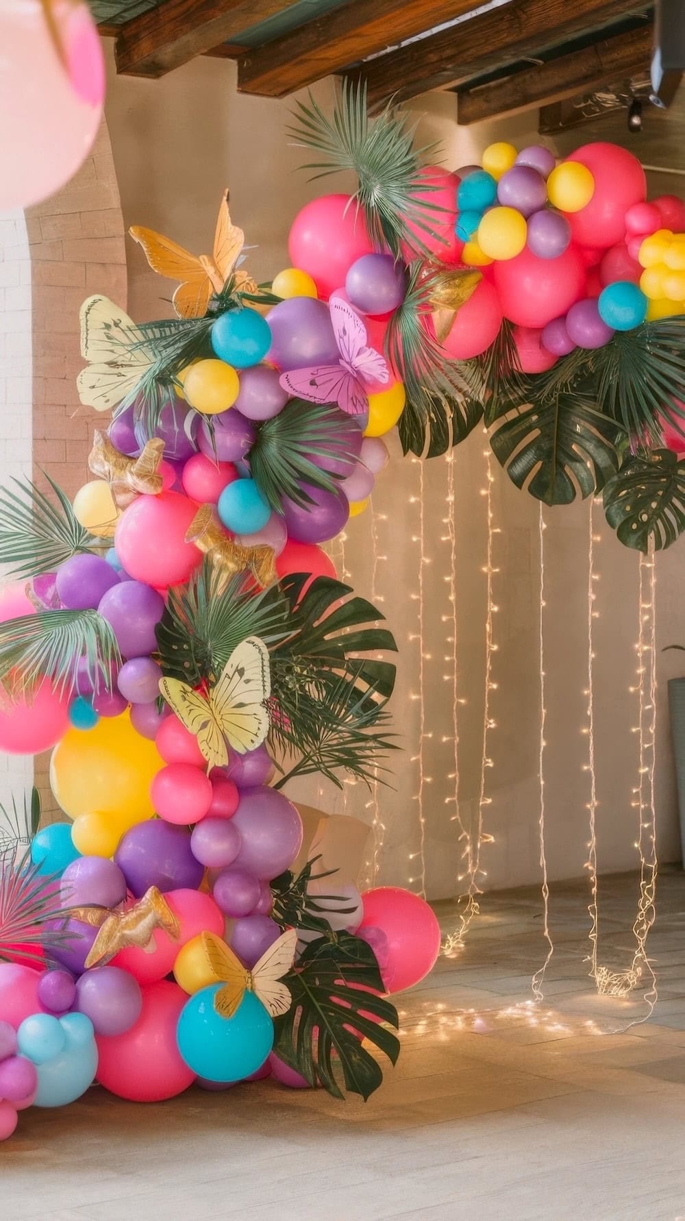 A vibrant balloon arch, perfect for butterfly baby shower ideas, features a mix of pink, purple, yellow, and blue balloons adorned with green leaves and decorative butterflies. String lights hang beneath the arch, creating a festive and colorful atmosphere.