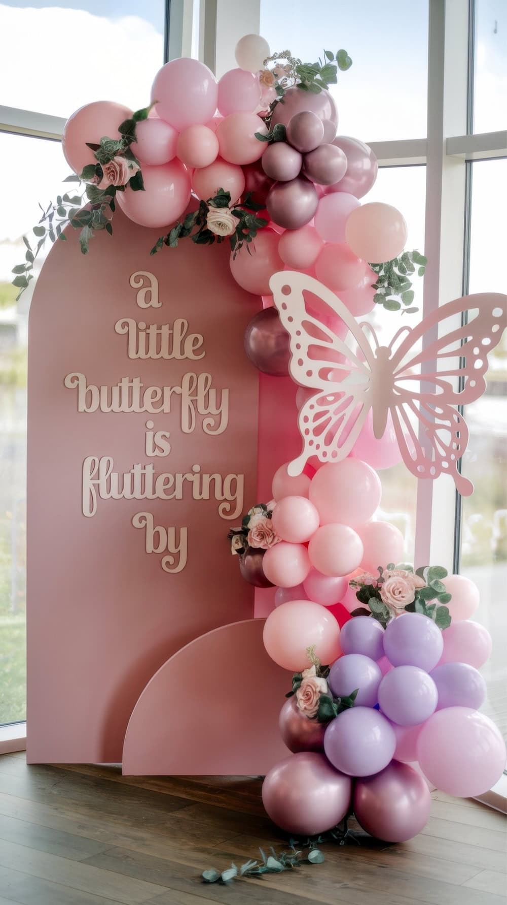 A pink and purple balloon arch adorned with flowers and a large butterfly cutout decorates the window, setting the stage for a whimsical butterfly baby shower. The sign reads, "a little butterfly is fluttering by," capturing the event's enchanting theme.