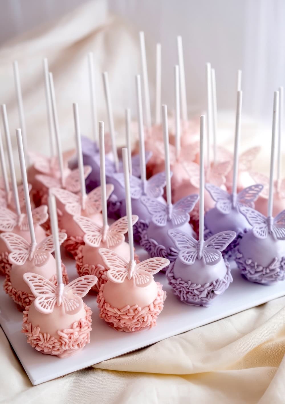 A tray of decorated cake pops with butterfly toppers showcases a delightful butterfly baby shower theme. Coated in pink and purple frosting, each pop features intricately detailed butterflies, neatly arranged on a white surface.