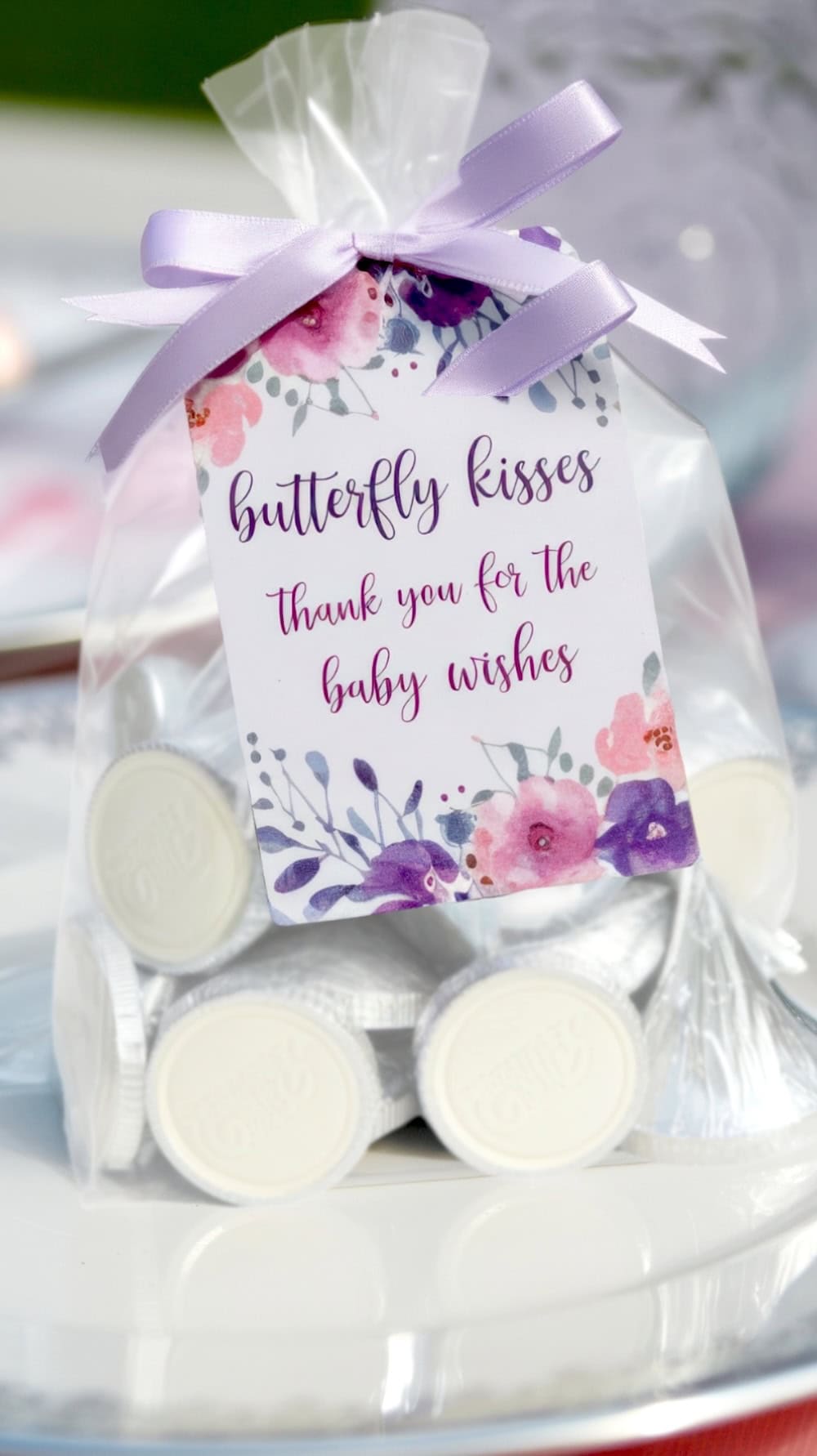 A clear bag tied with a lavender ribbon holds white candies, perfect for a butterfly baby shower. Attached is a floral card adorned with purple and pink flowers that reads, "butterfly kisses, thank you for the baby wishes.
