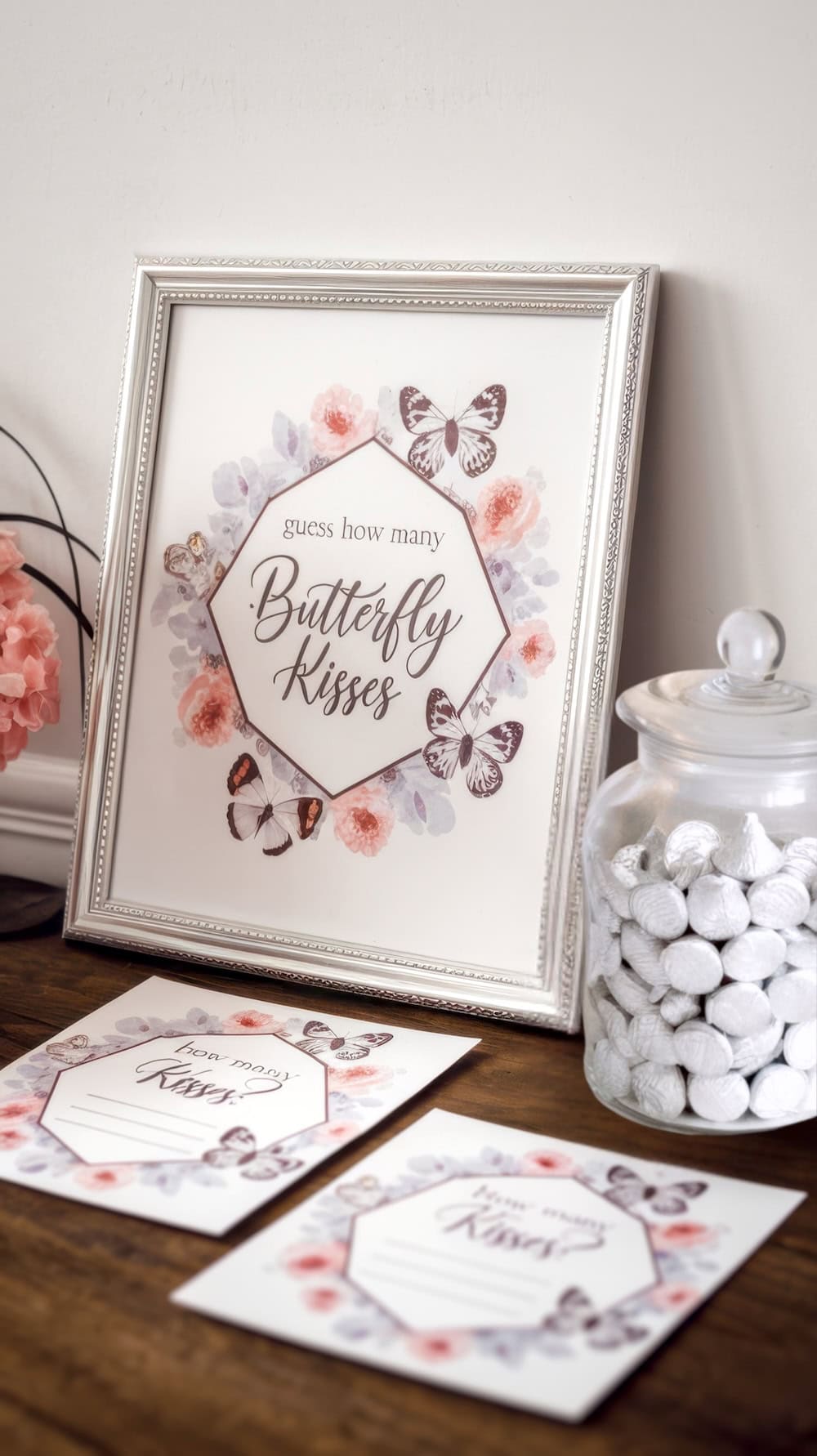 A framed sign with "guess how many Butterfly Kisses" sits inside a floral and butterfly border, perfect for a butterfly baby shower. Nearby, an open jar brims with white candies, while matching design cards rest on a wooden surface. A pink flower blooms in the background.