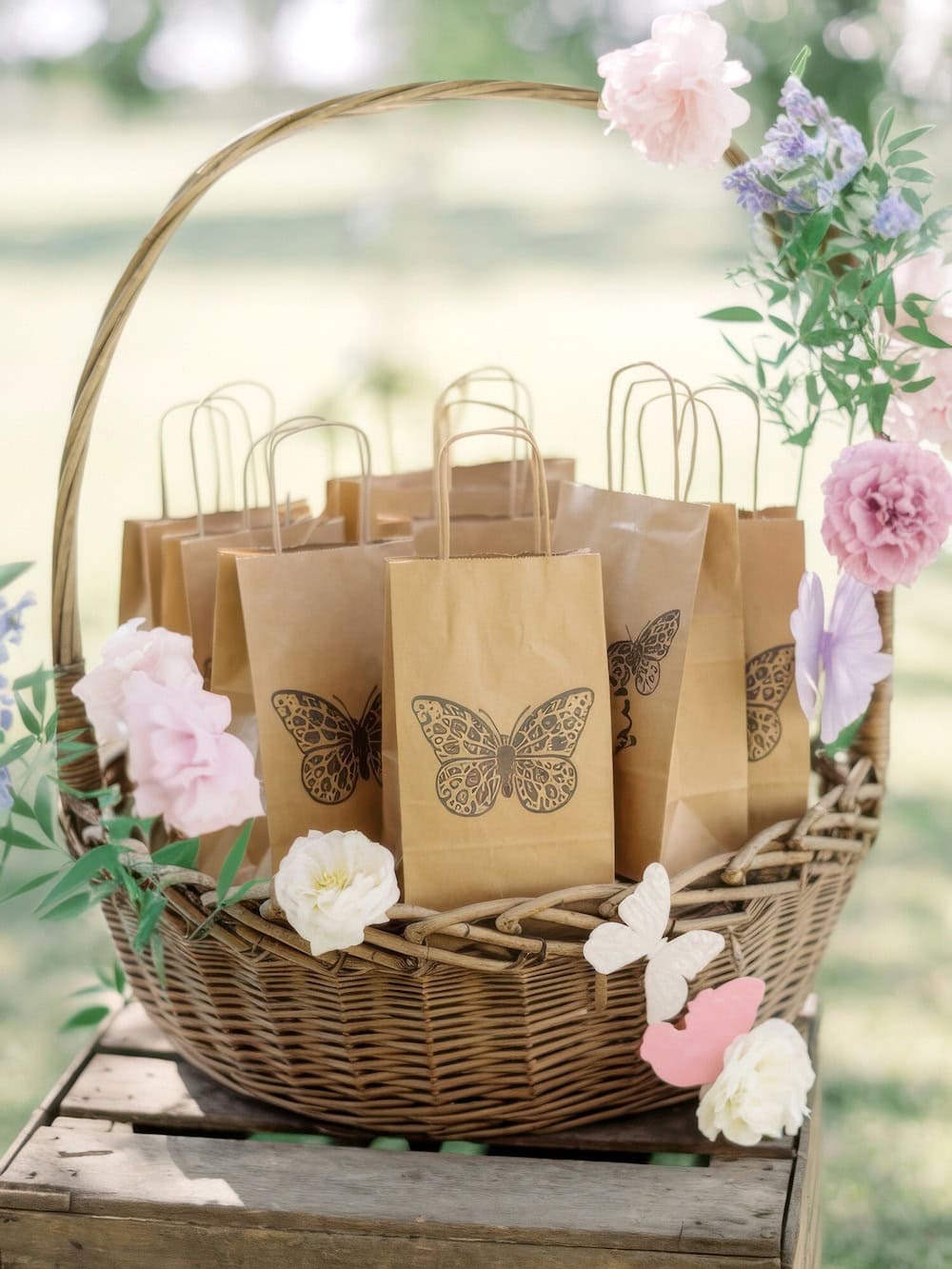 A wicker basket brimming with brown paper bags featuring butterfly designs is perfect for a butterfly baby shower. Adorned with pink and white flowers, it sits outdoors on a wooden surface against a soft-focus green background.