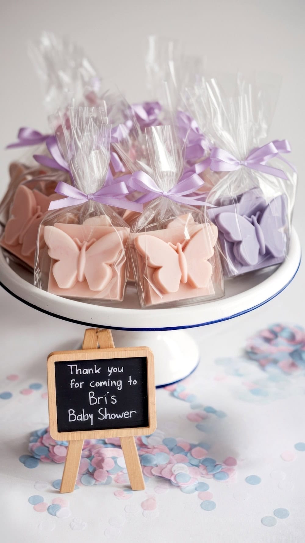 A white cake stand holds butterfly baby shower party favors or prizes with butterfly-shaped soaps in pink and purple, each tied with a lilac ribbon. In front, a small chalkboard sign reads, "Thank you for coming to Bri's Baby Shower." Confetti is scattered on the table.