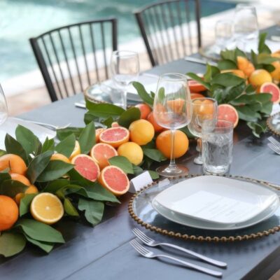 An elegant outdoor table setting perfect for little cutie baby shower ideas, featuring a citrus-themed centerpiece with oranges, grapefruits, and leaves. Clear plates with gold-rimmed chargers and glassware complete the look, set against a softly blurred pool in the background.