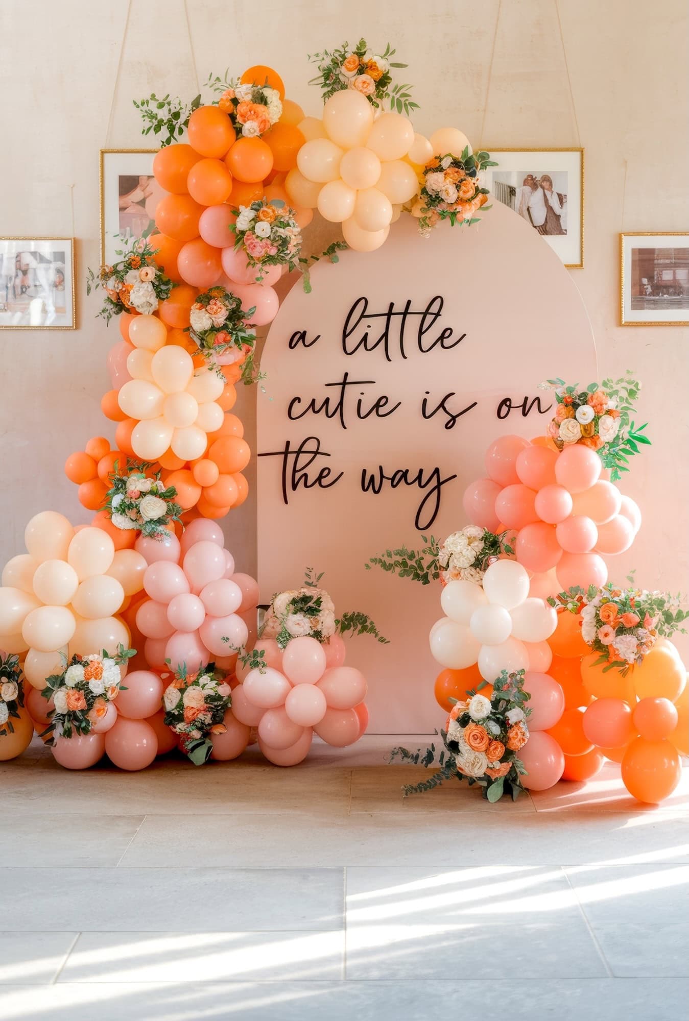 A balloon arrangement in shades of orange, peach, and pink surrounds a sign that reads "a little cutie is on the way," perfect for baby shower ideas. The setup includes flowers and greenery, with framed photos on the wall behind.