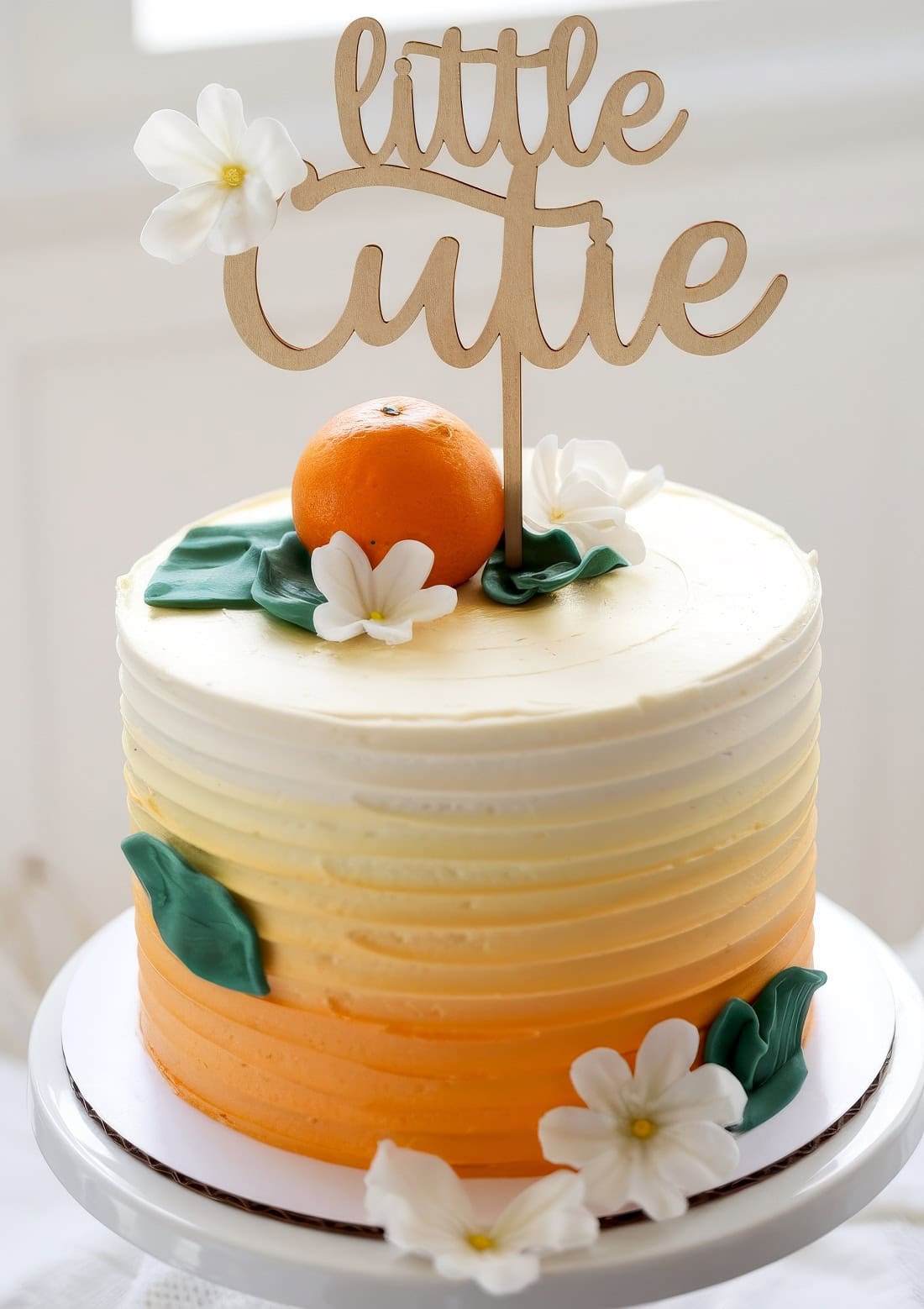 A round cake with gradient layers of white to orange icing is perfect for little cutie baby shower ideas. It is adorned with white flowers, green leaves, and a small orange. A wooden topper reads "little cutie." The cake sits on a white stand against a bright background.