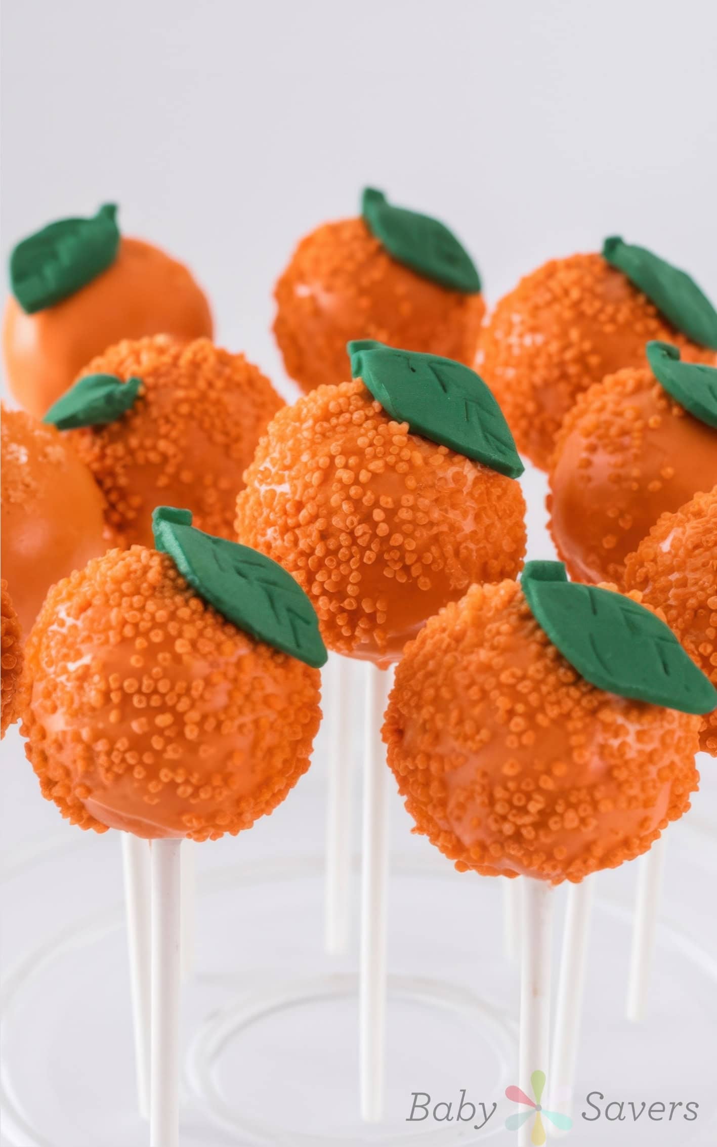 Orange cake pops resembling small pumpkins, each topped with a green fondant leaf, create the perfect dessert to add to your listof little cutie baby shower ideas. They are set on white sticks against a plain background.