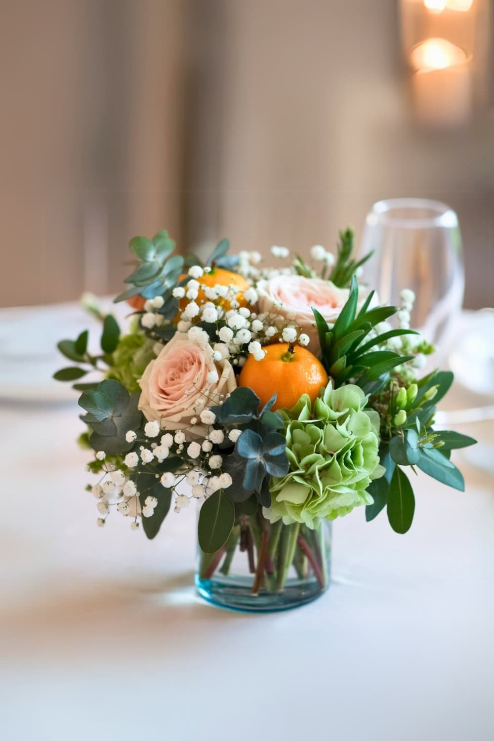 A vase centerpiece for a little cutie baby shower features light pink roses, orange fruits, green hydrangeas, eucalyptus leaves, and white baby's breath. It rests on a white tablecloth with blurred glassware and soft lighting in the background, creating a charming and delicate ambiance.