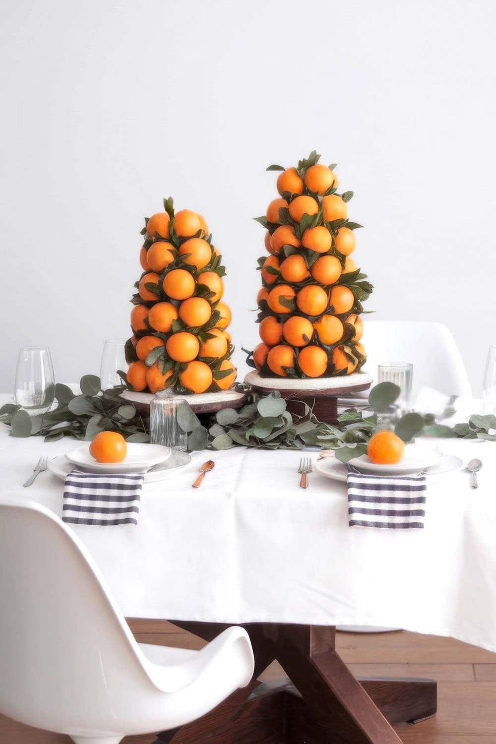 Two cone-shaped towers made of clementine oranges and green leaves are on a table with a white tablecloth, perfect for delightful little cutie baby shower ideas. The table is set with plates, black and white checkered napkins, and glassware. Eucalyptus branches run across the center. White chairs surround the table.