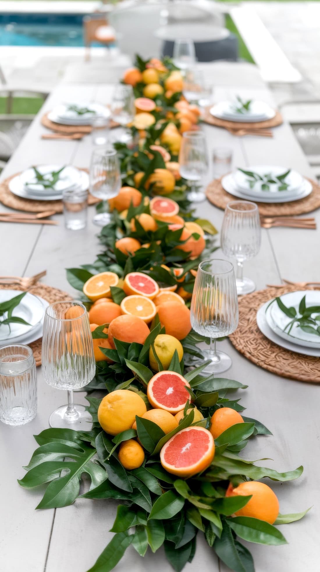 A long table is elegantly set with plates, glasses, and woven chargers. A lush centerpiece of assorted citrus fruits and leafy greens runs down the middle, echoing little cutie baby shower ideas in this charming outdoor setting. A pool glistens in the background.
