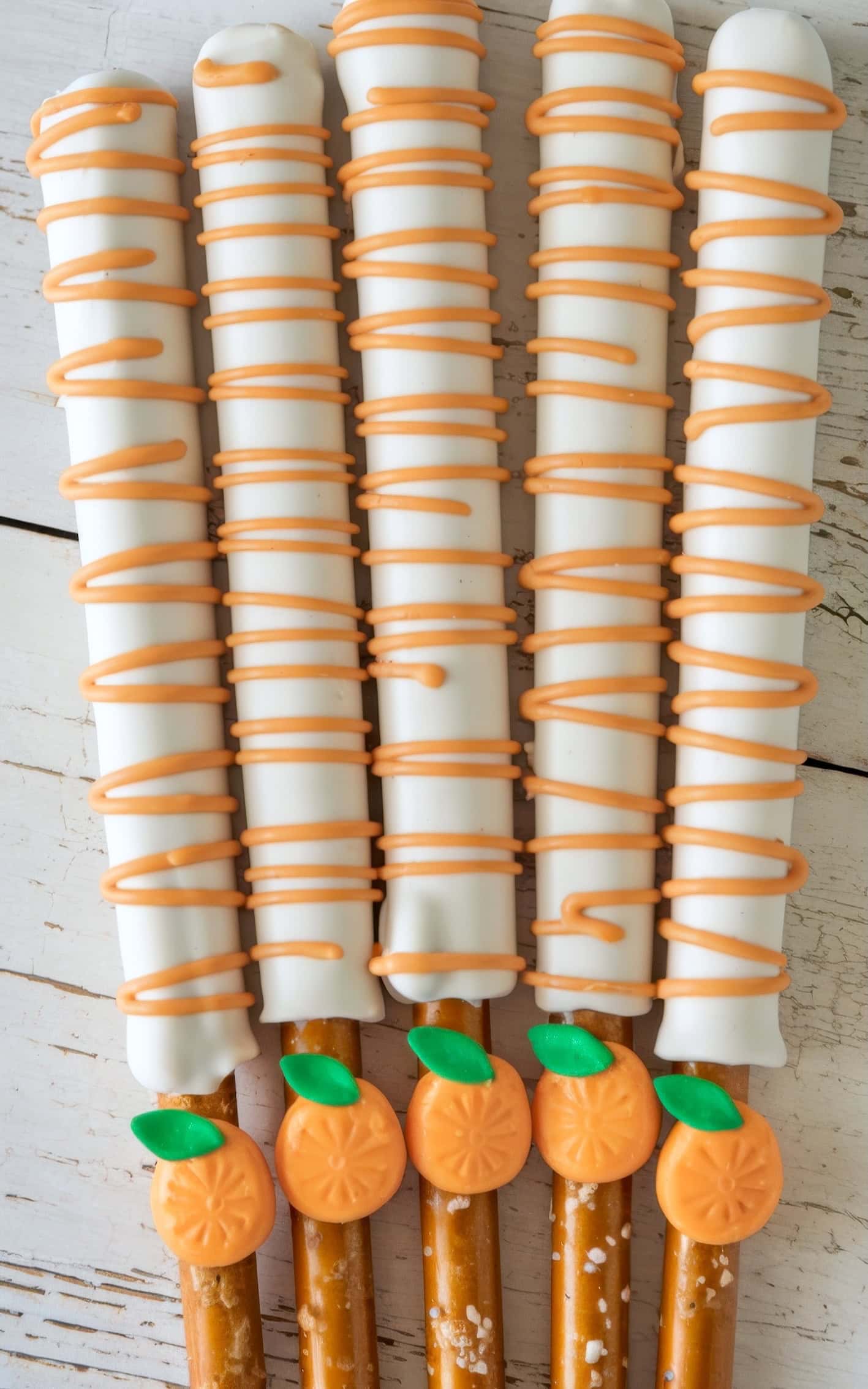 Five pretzel rods are coated in white chocolate and drizzled with orange icing, each adorned with an orange citrus decoration. Perfect for little cutie baby shower ideas, they are arranged in a row on a white wooden surface, offering a charming treat for celebrations.