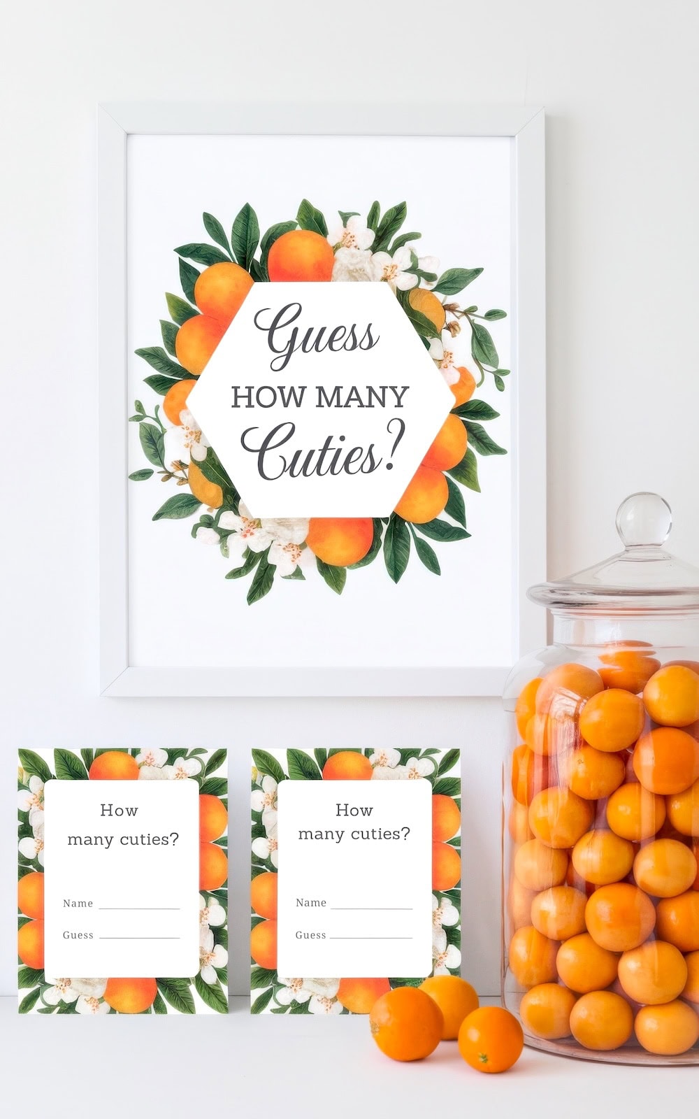 Framed sign with floral design and text "Guess How Many Cuties?" Two cards below ask, "How many cuties?" with space for name and guess—a charming baby shower idea. A jar filled with little orange cutie fruits is on the right.