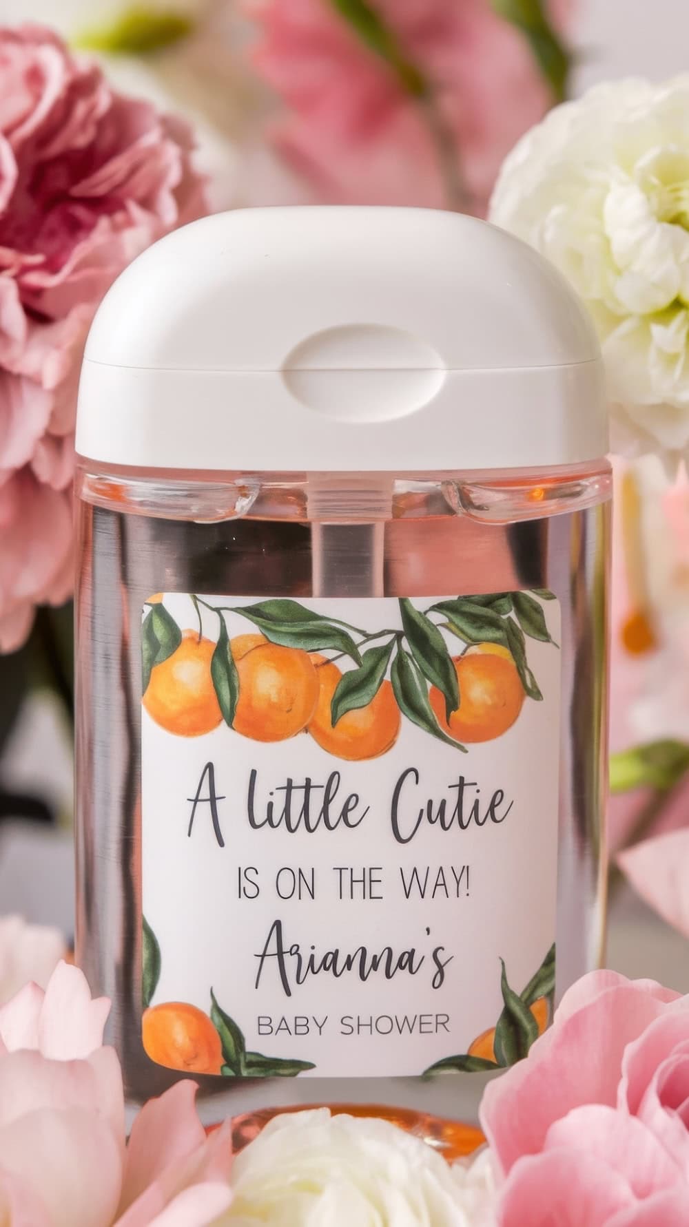 A clear bottle with a white lid is nestled among pink and white flowers. The label, adorned with orange illustrations, reads, "A little cutie is on the way! Arianna's Baby Shower." This charming detail adds a sweet touch to your baby shower ideas.