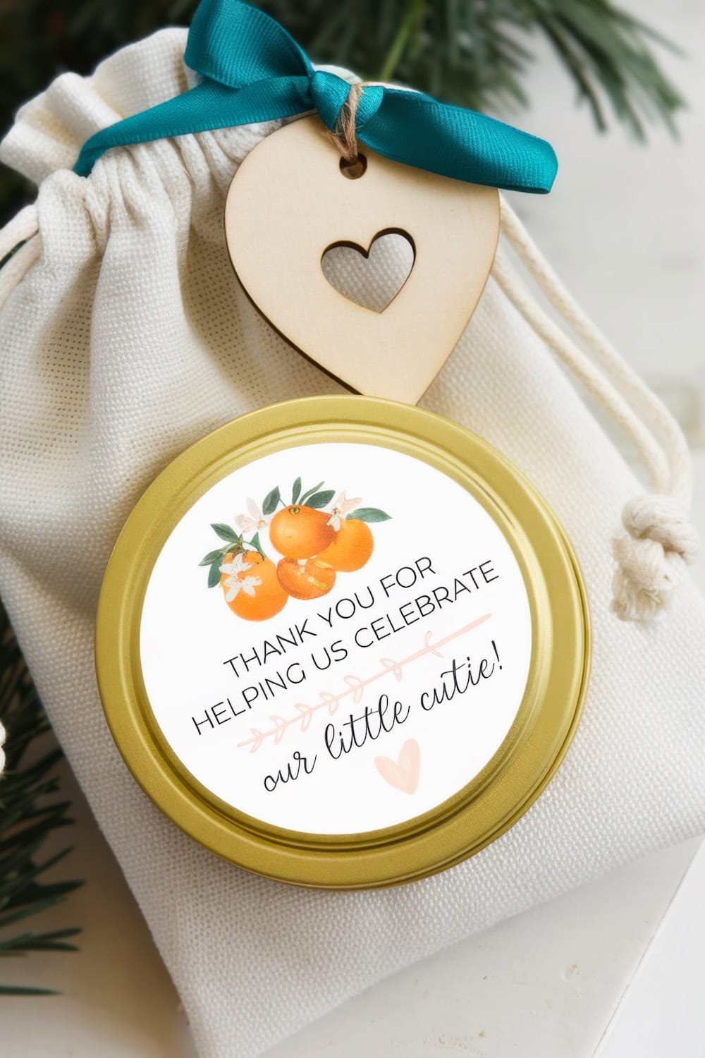 A small white drawstring bag with a wooden heart tag contains a jar with a golden lid, perfect for little cutie baby shower ideas. The label features oranges and flowers, reading, "THANK YOU FOR HELPING US CELEBRATE our little cutie!" The scene is complete with pine branches.