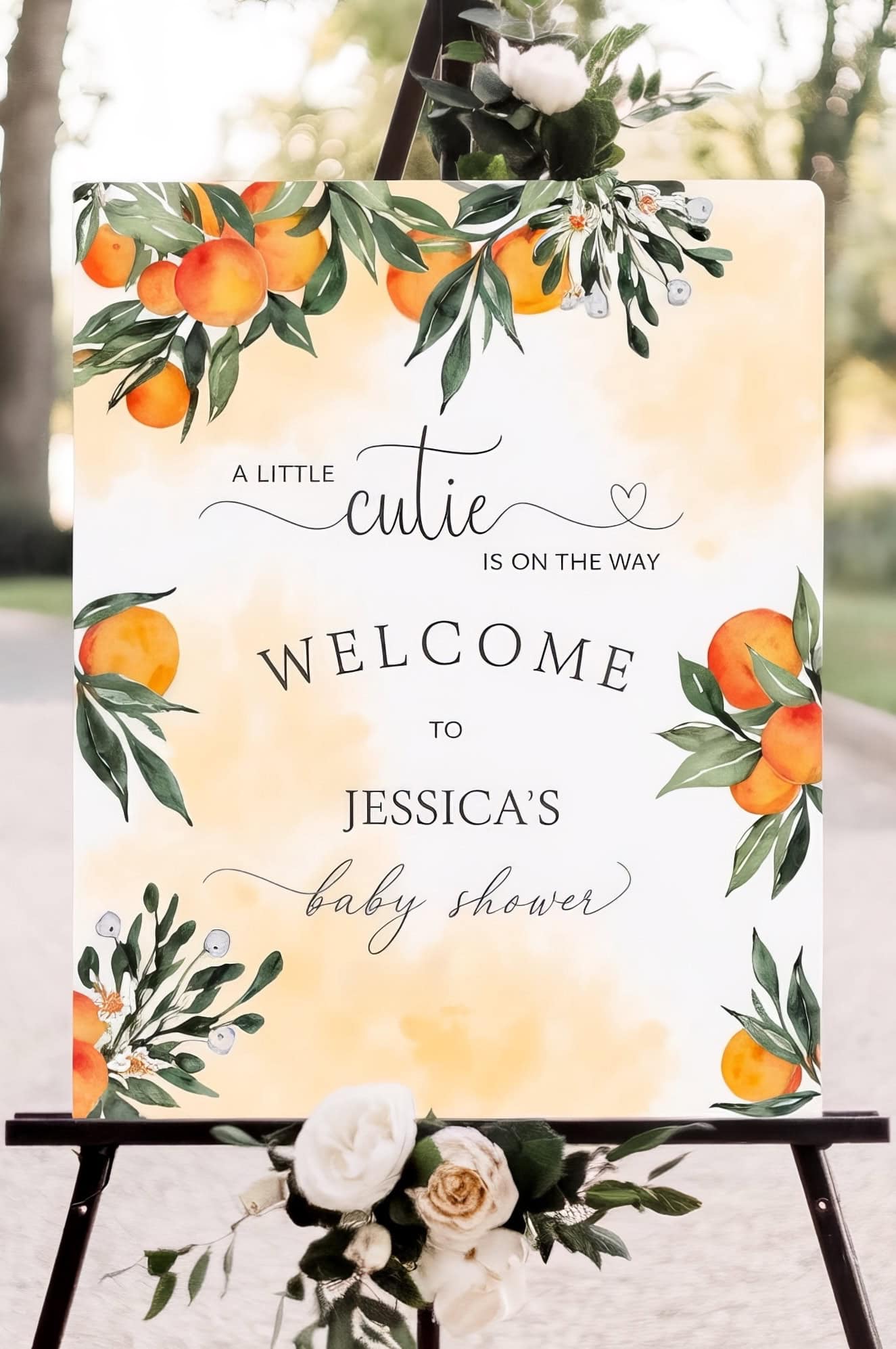 A decorative sign on an easel showcases orange fruit and greenery illustrations, reading: “A little cutie is on the way. Welcome to Jessica’s baby shower.” Florals adorn the base, offering charming little cutie baby shower ideas for your celebration.