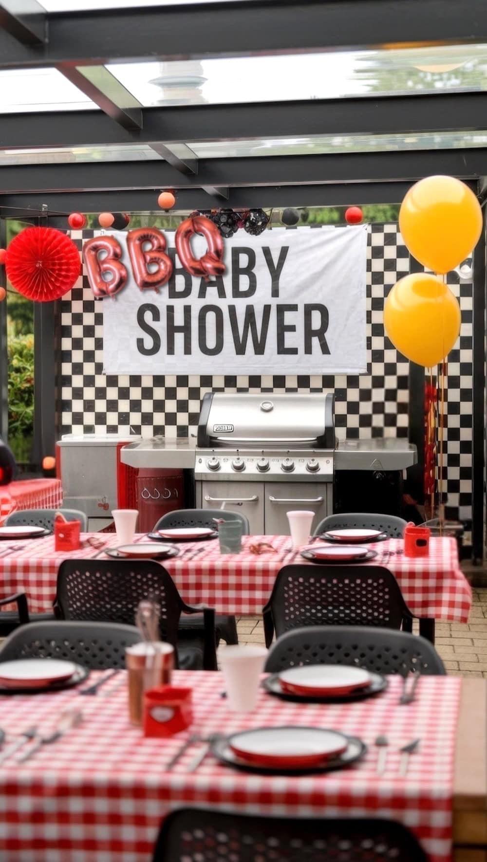 The BBQ-themed baby shower setup features a checkered backdrop with a lively BBQ BABY SHOWER banner. Tables are adorned with red and white checkered cloths, complemented by yellow balloons, all set against the backdrop of a large barbecue grill.