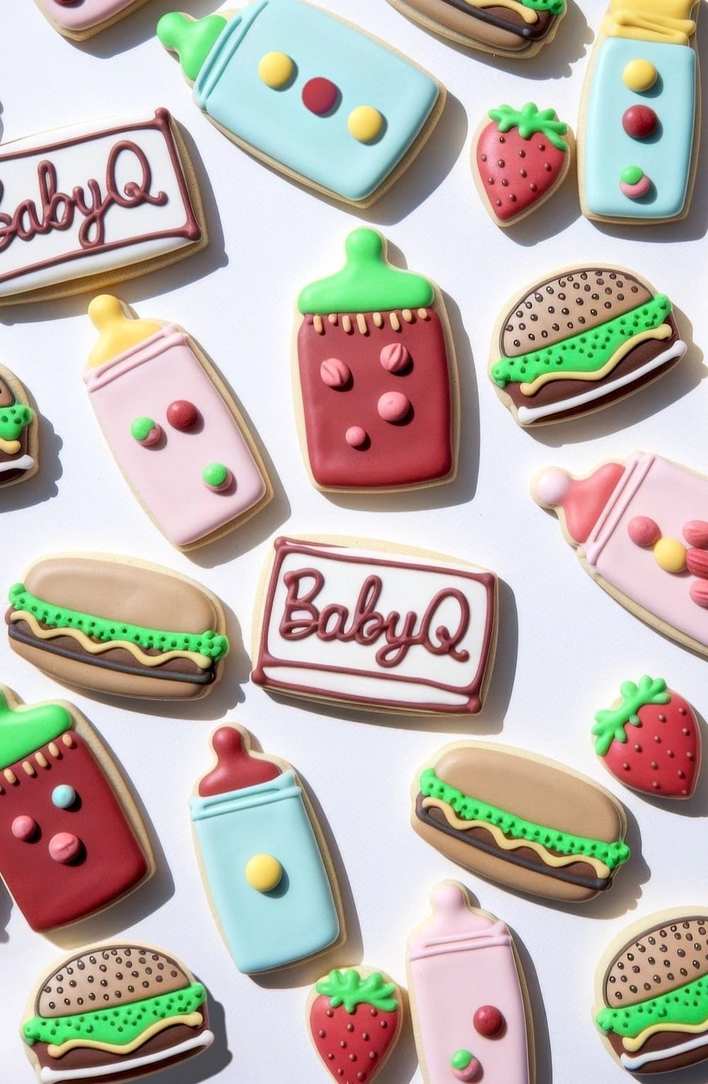 A variety of colorful, decorated cookies in shapes like baby bottles, strawberries, hamburgers, and hot dogs are arranged on a white background. Perfect for a BBQ baby shower theme, some cookies even have "BabyQ" written on them. The icing is bright and detailed.