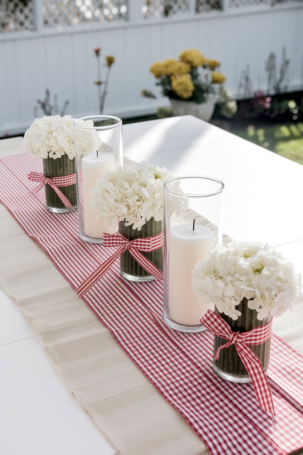 A table centerpiece perfect for a BBQ baby shower features alternating clear glass cylinders of white hydrangeas and pillar candles, each tied with red checkered ribbons, all beautifully arranged on a red and white checkered runner atop a crisp white table.