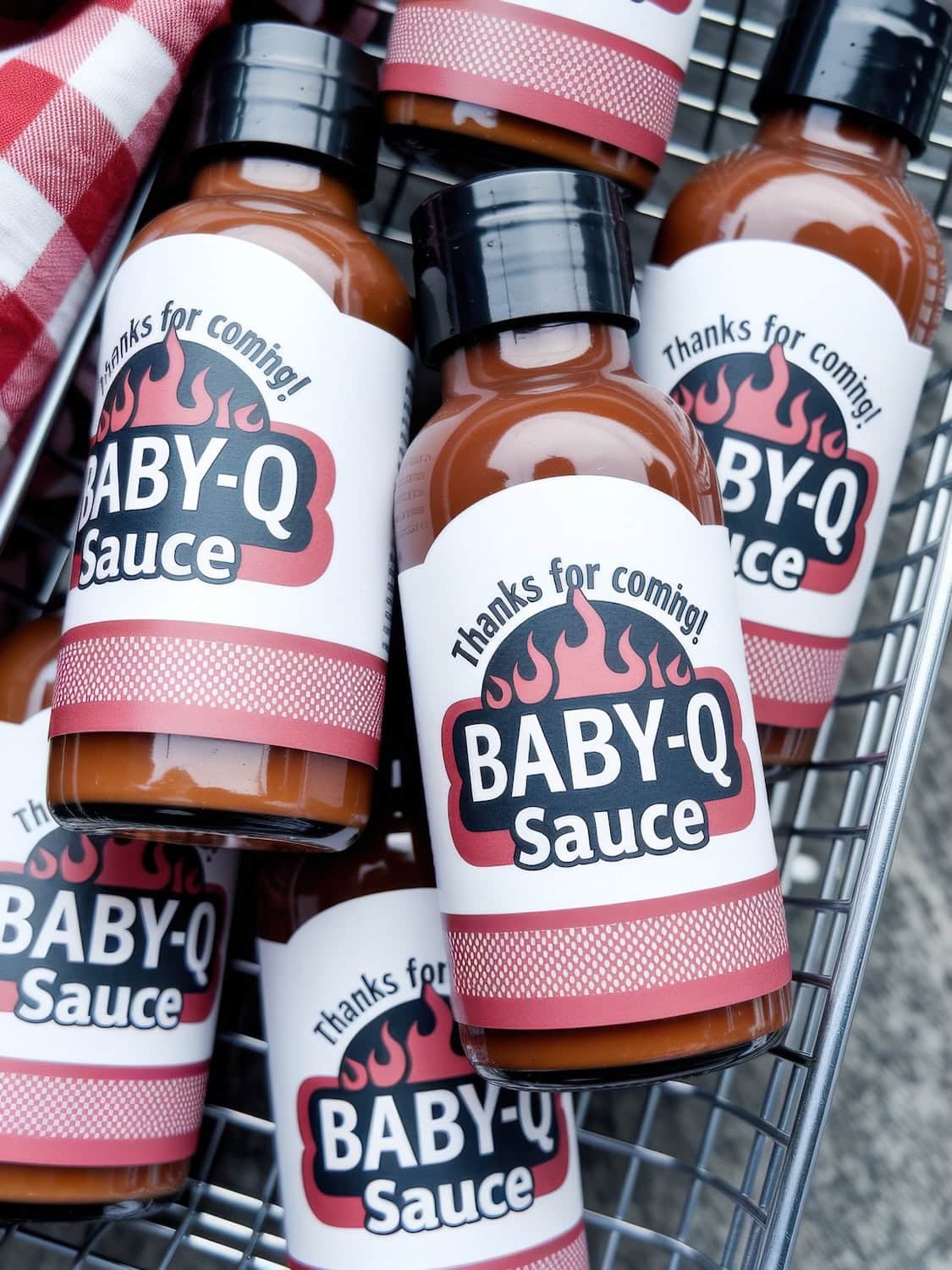 Several bottles of "Baby-Q Sauce" with the label "Thanks for coming!" are displayed in a metal basket, perfect for a BBQ baby shower. The sauce, playfully named, adds charm to any barbecue event. A red and white cloth is partially visible.