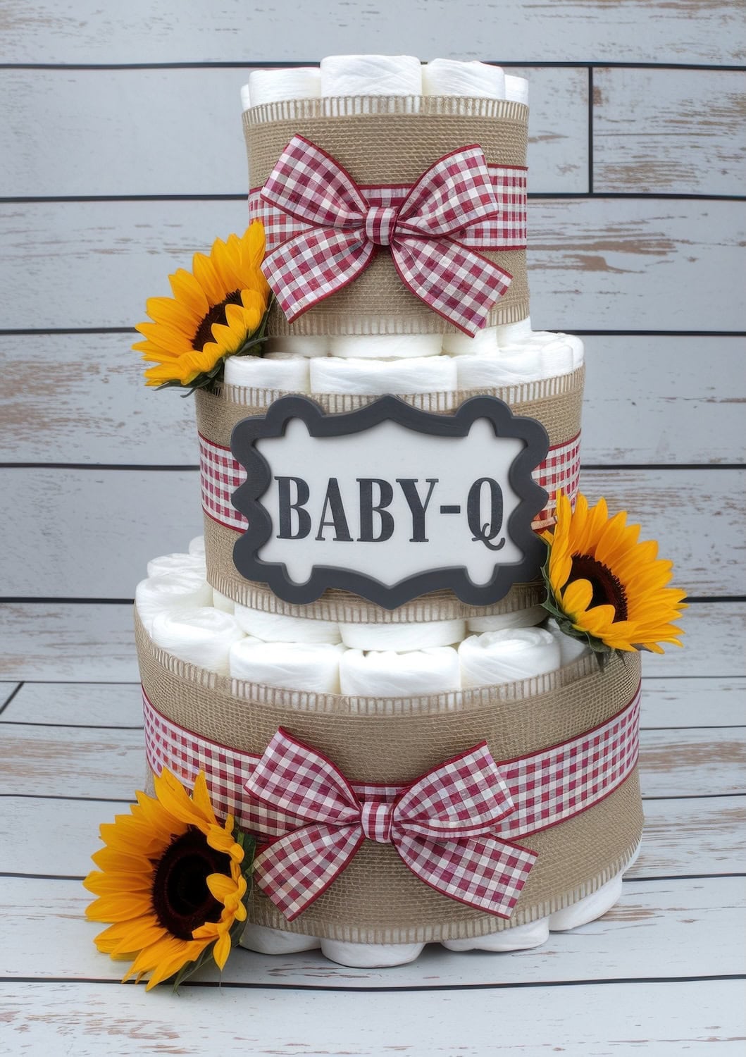 A three-tier diaper cake, perfect for a BBQ baby shower, is decorated with red-and-white gingham ribbons, burlap, and sunflowers. A sign on the middle tier reads "BABY-Q," set against a white wooden background.